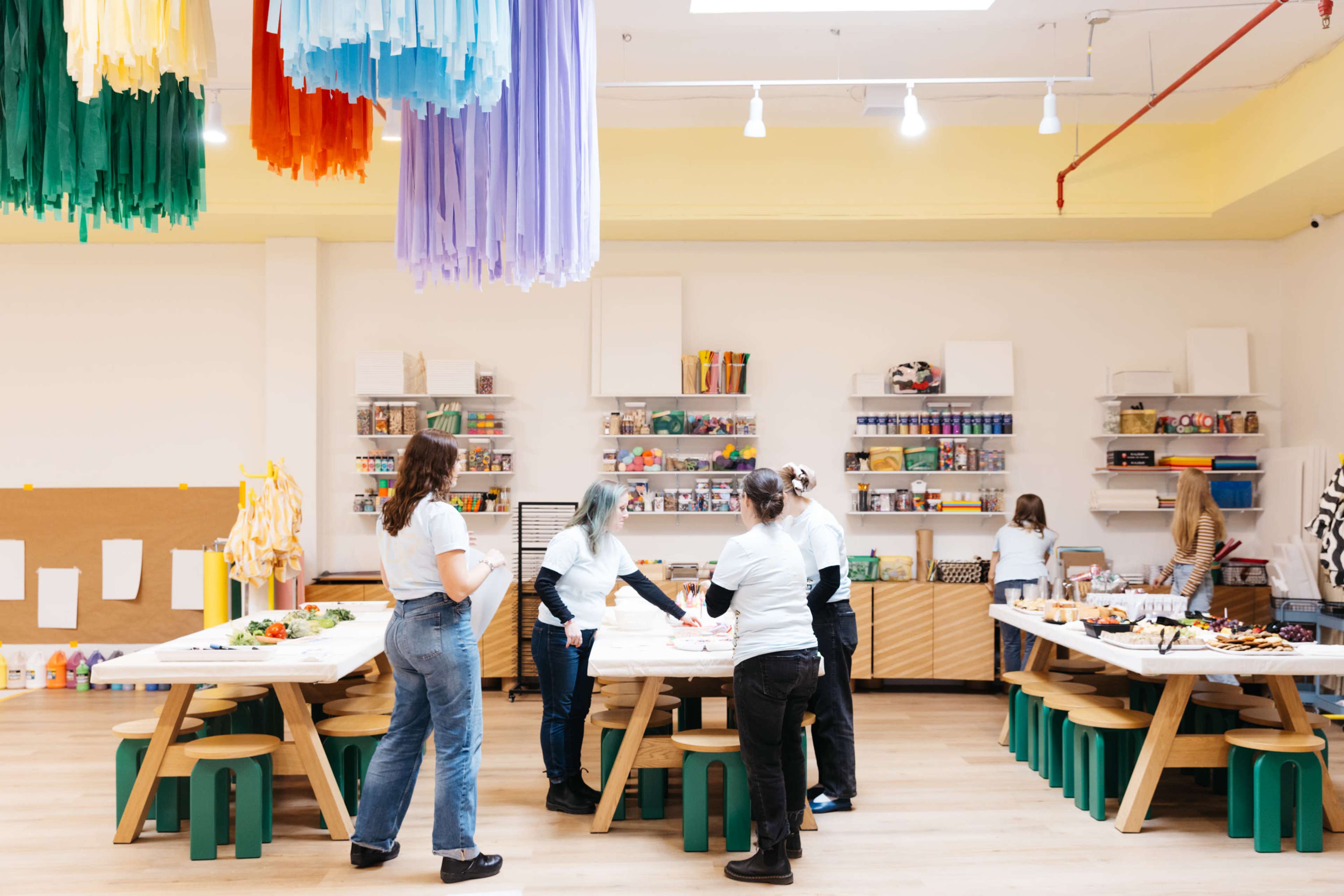 A group of people are collaborating around large tables in a bright, organized creative workspace adorned with colorful paper decorations.