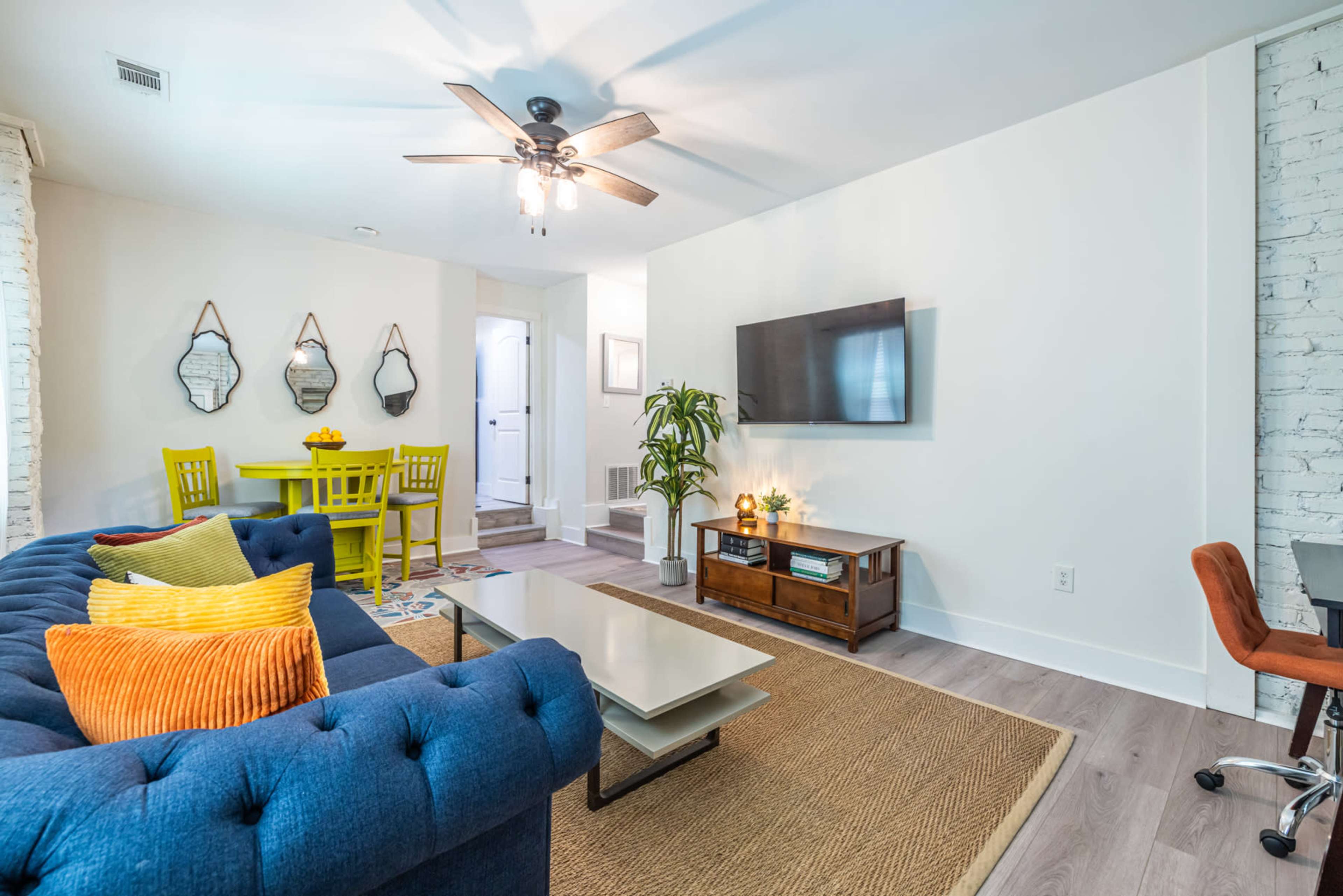 The image shows a modern living room with a blue sofa, a wooden coffee table, a flat-screen TV on the wall, and a yellow dining set in the background.
