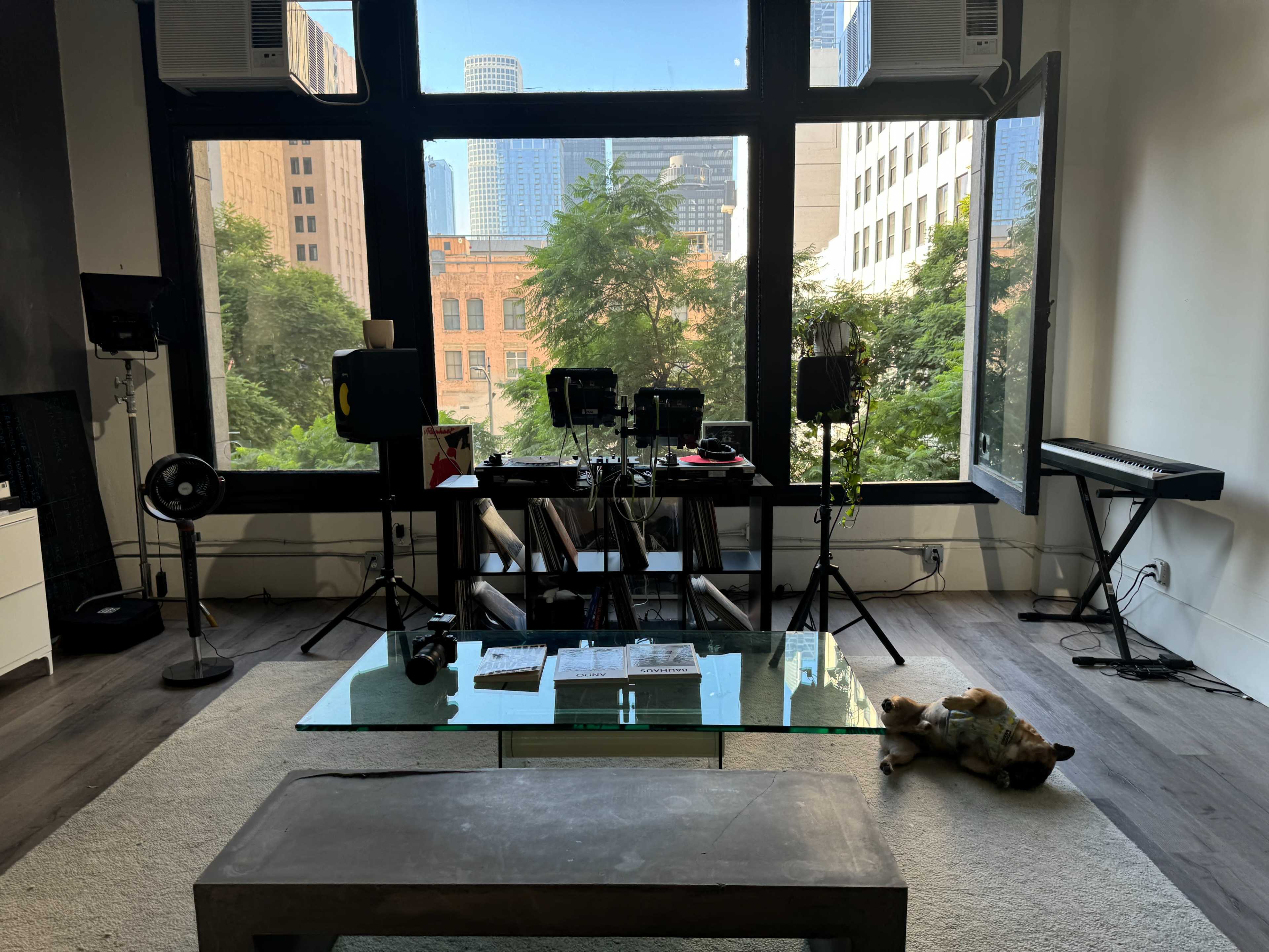 The image shows a modern room with large windows overlooking a cityscape, featuring a glass coffee table, a few electronic devices, and a plush toy on the floor.