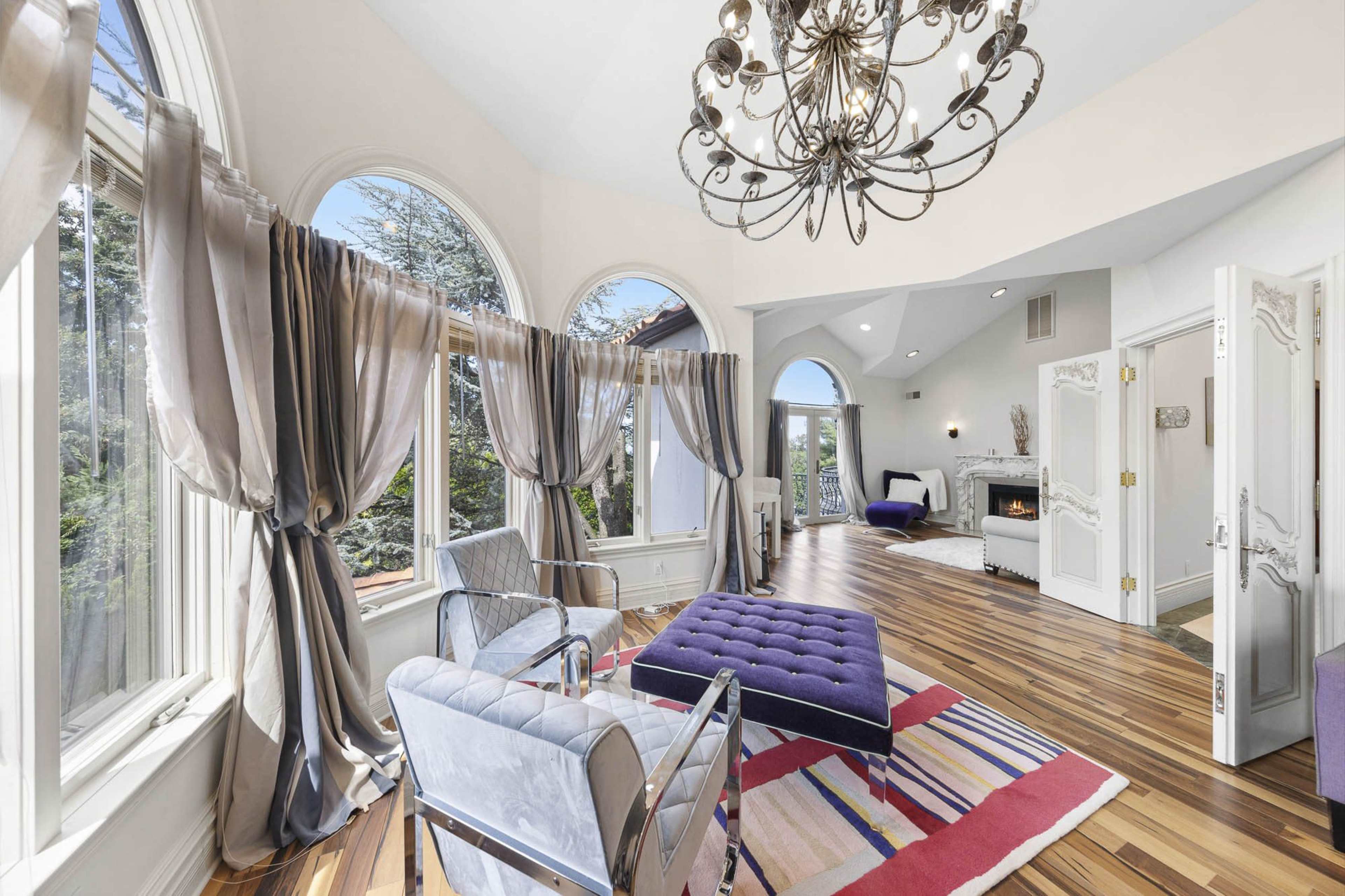 The room features large arched windows, elegant drapes, a purple upholstered ottoman, and wooden flooring with a variety of colorful rugs.