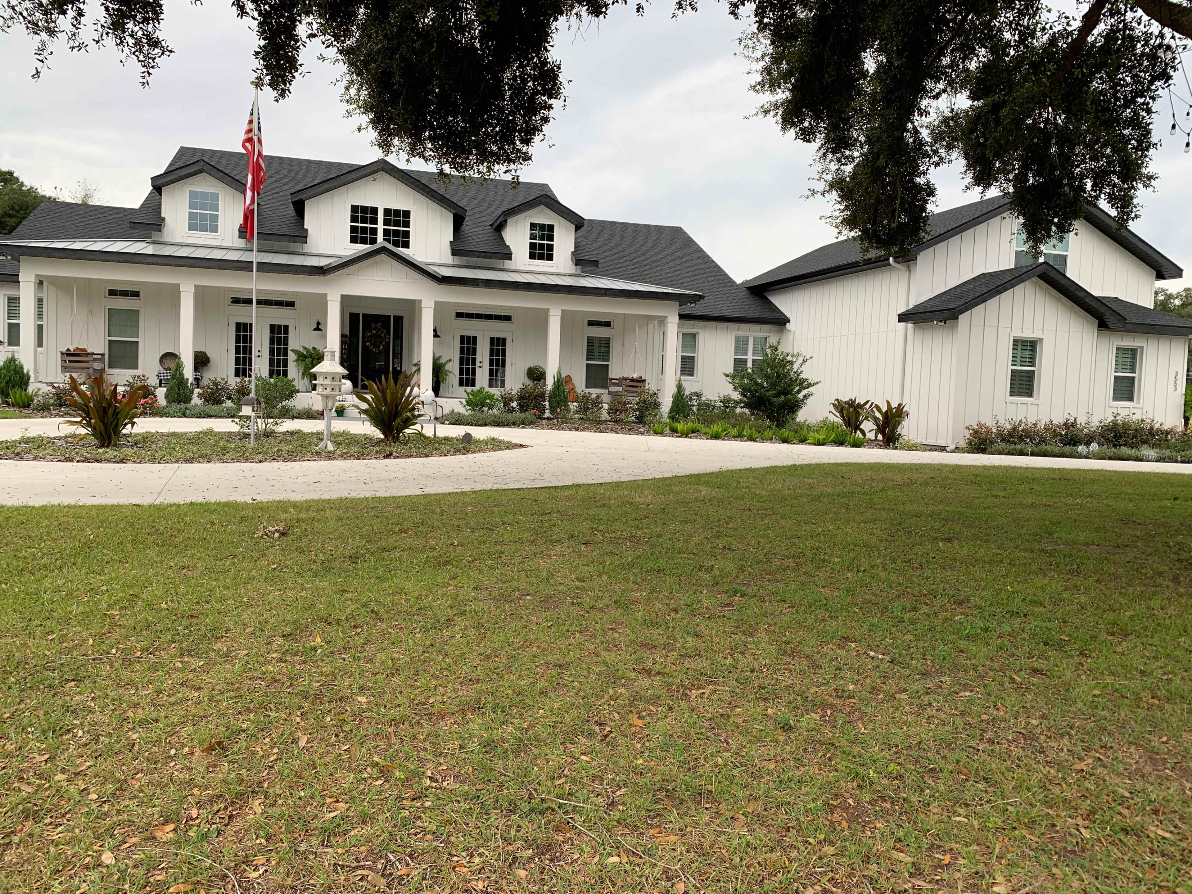 The image shows a large, two-story white house with a black roof, situated on a well-maintained lawn, and featuring a flagpole at the front.