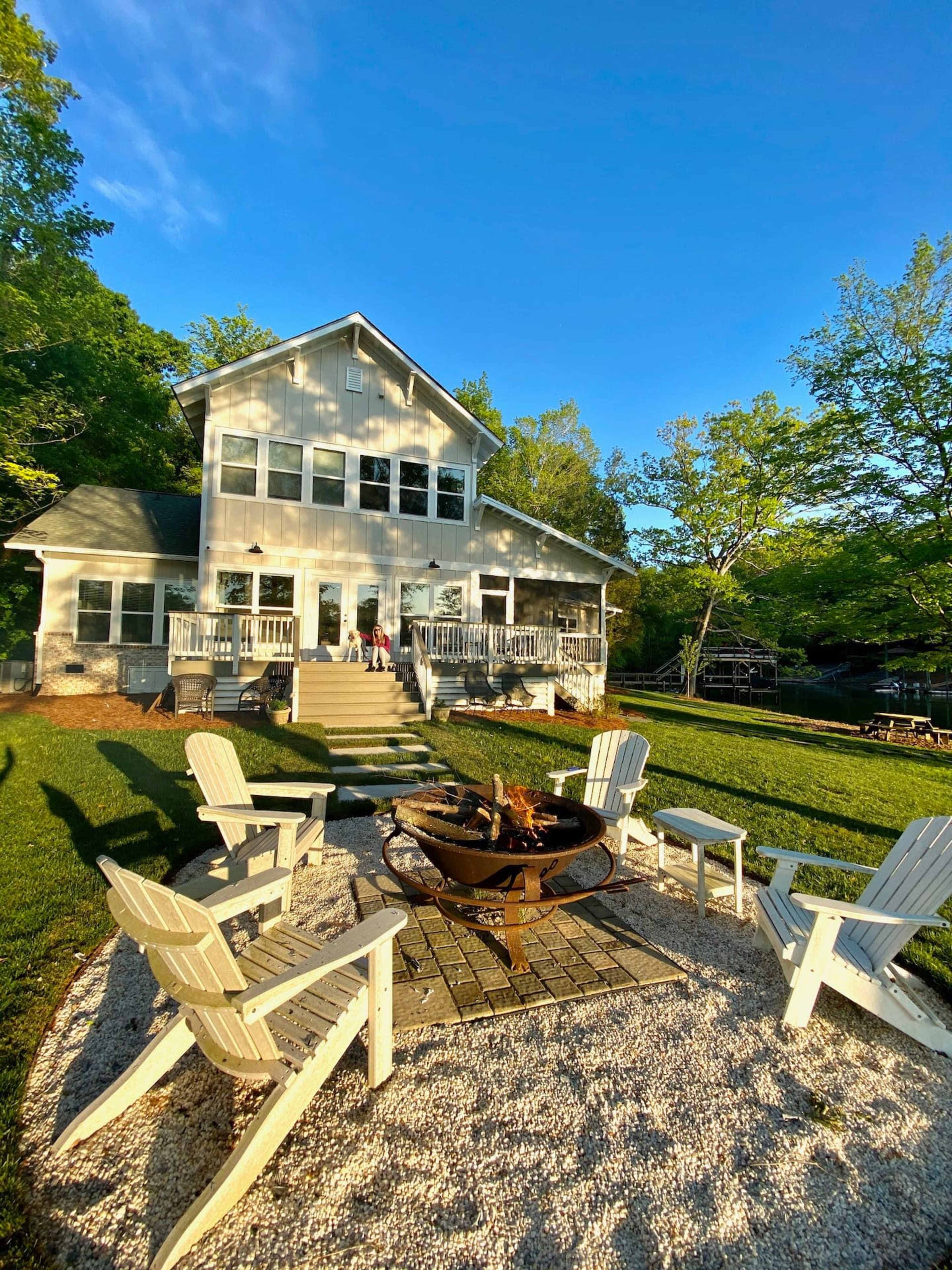 A modern two-story house with a spacious backyard features a fire pit surrounded by Adirondack chairs and lush greenery.