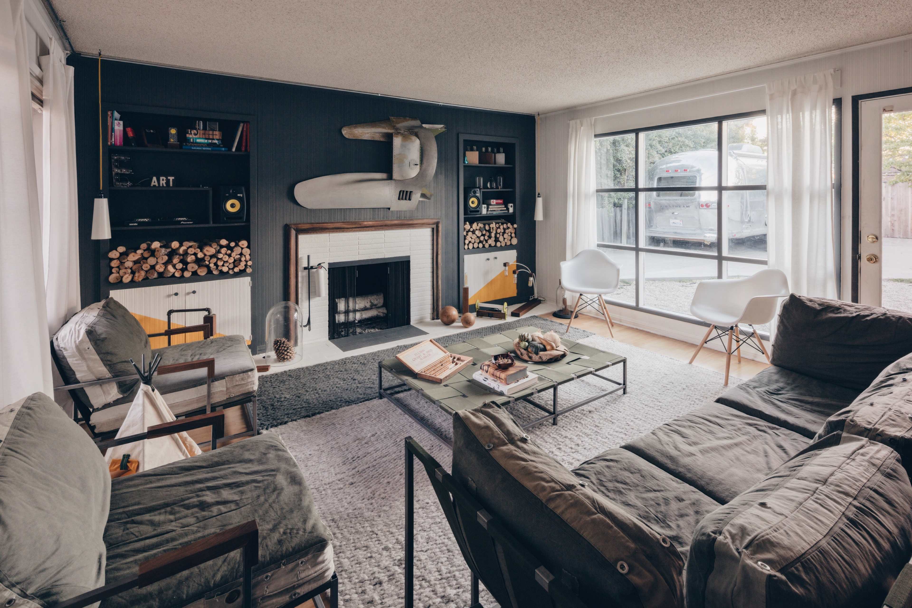 A cozy living room features a fireplace, stylish seating, and shelves with decorative items, all illuminated by natural light from a large window.