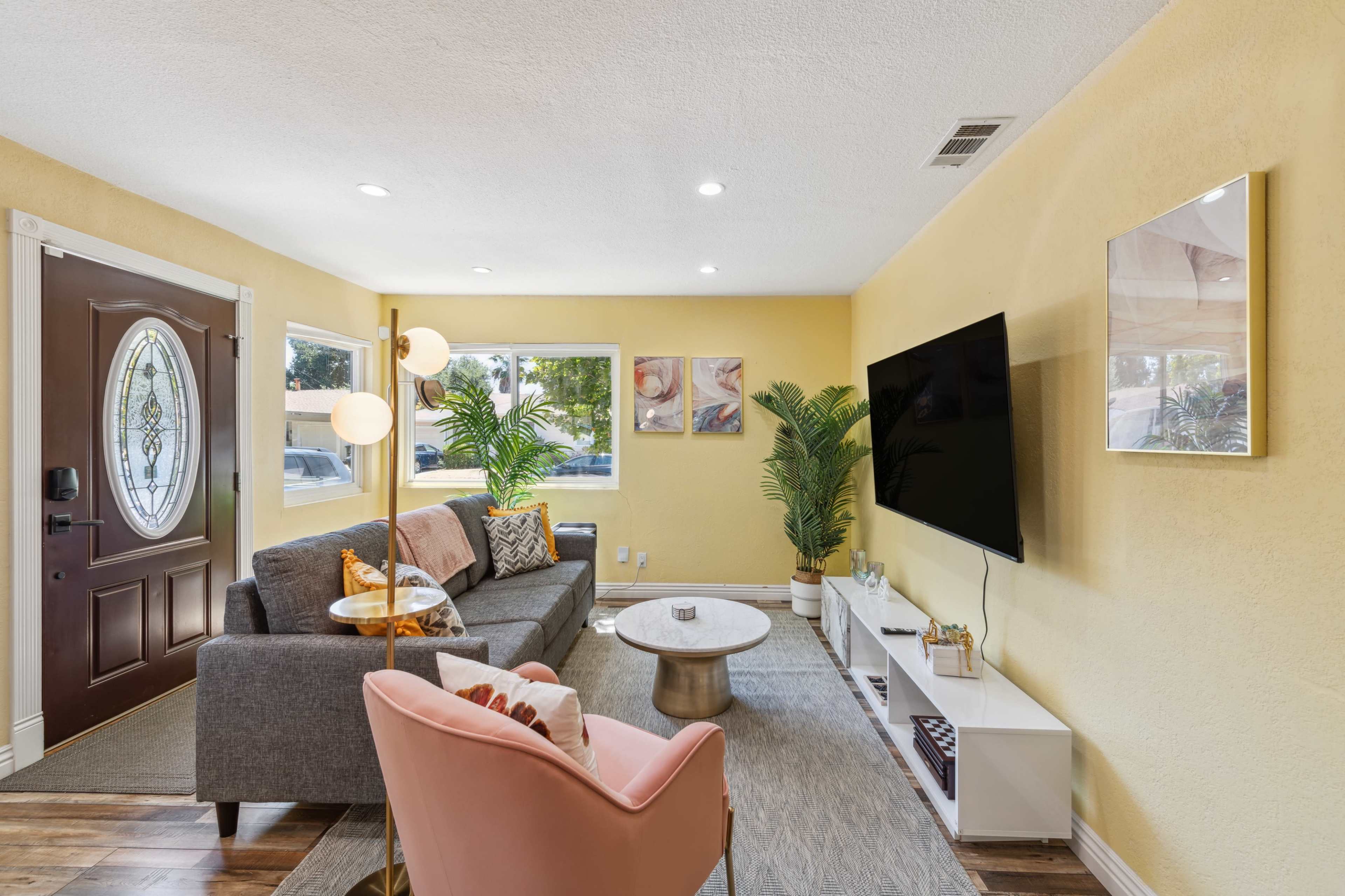 The living room features a gray sofa, a pink armchair, a circular coffee table, and a large TV mounted on a yellow wall with decorative plants and artwork.