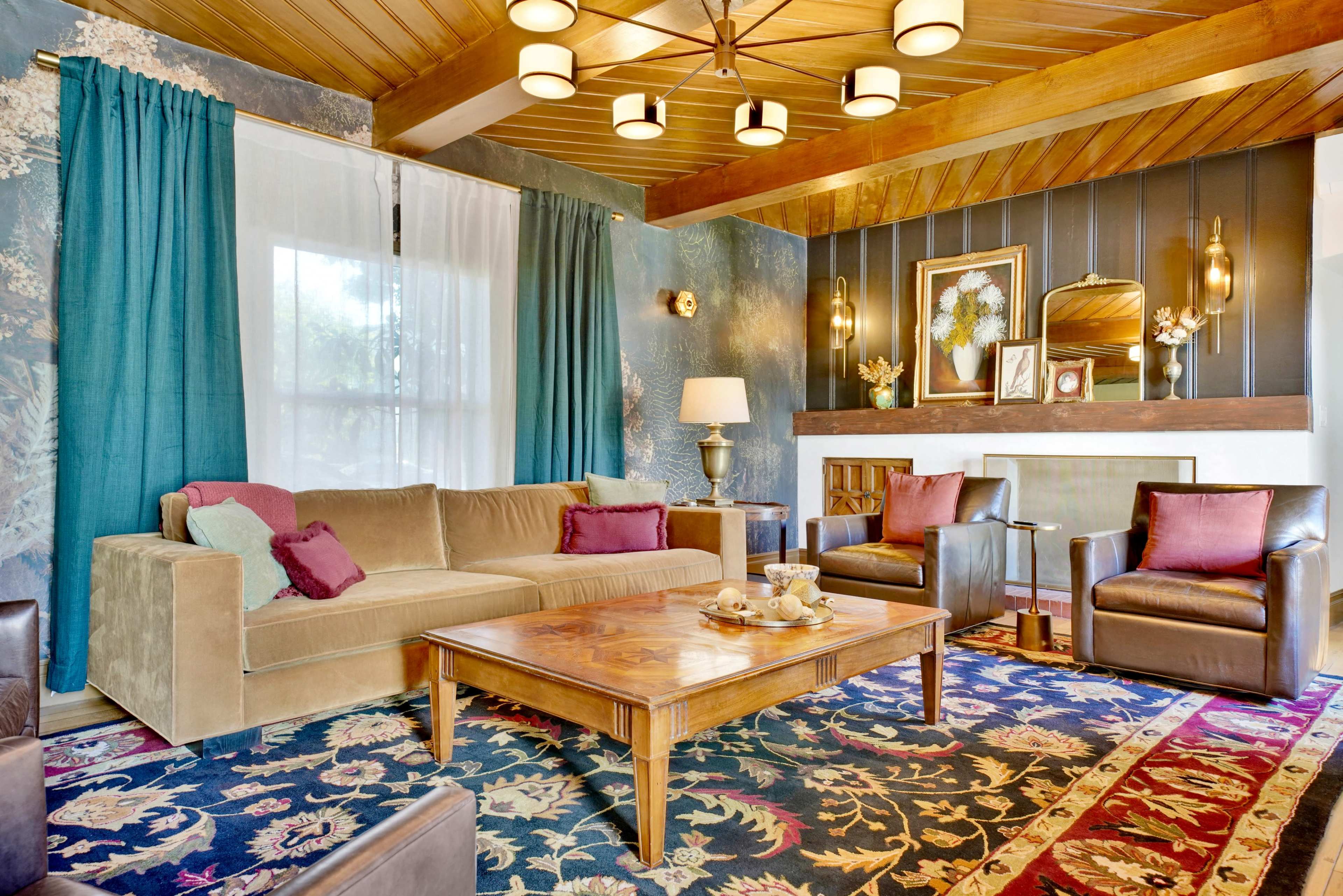 The image shows a stylish living room featuring a beige sofa, two dark leather armchairs, a wooden coffee table, and a colorful area rug, with large windows dressed in green curtains.