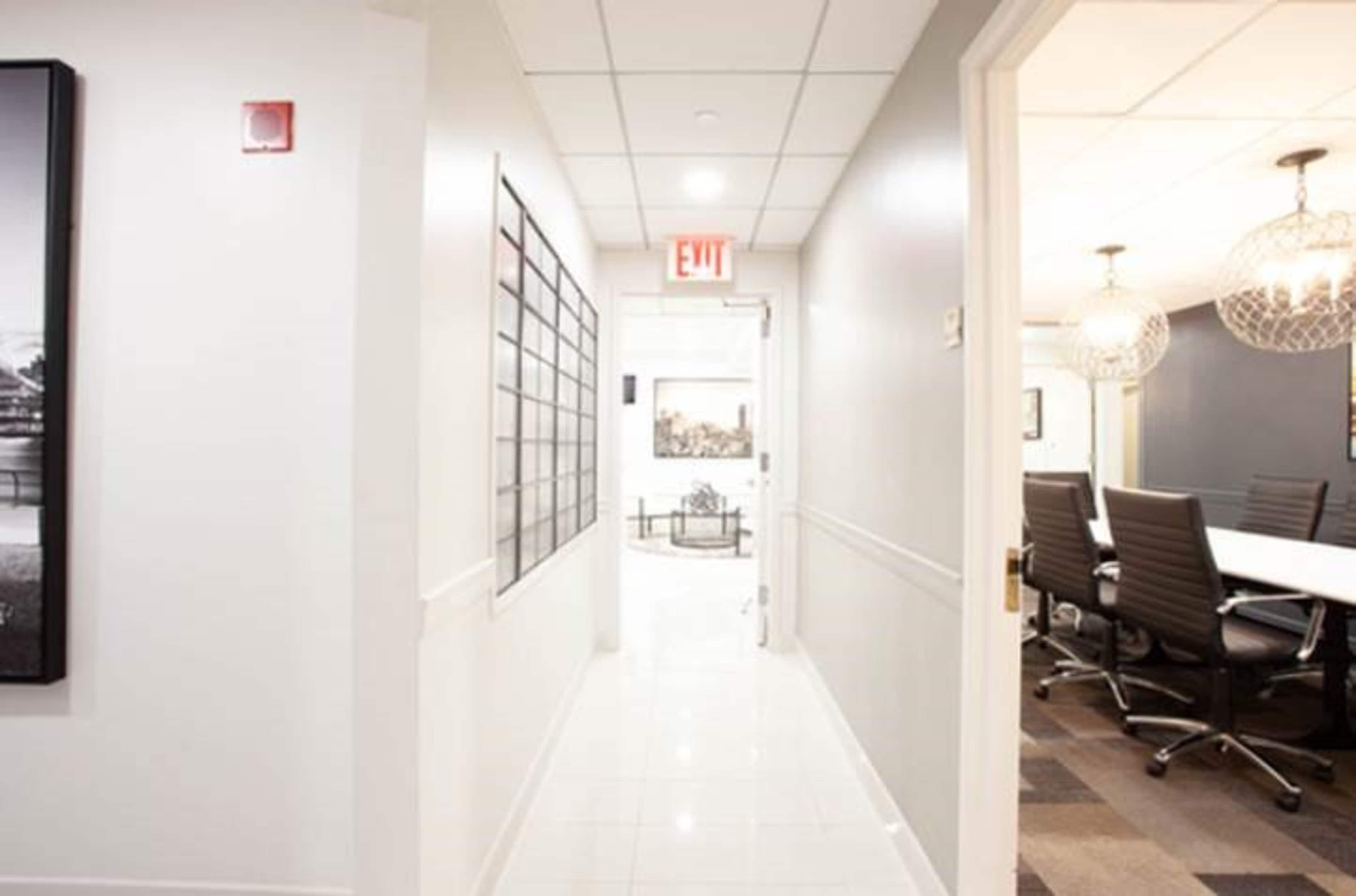 A brightly lit corridor with white walls and tiled flooring leads to an exit door on one end and a meeting room on the other.