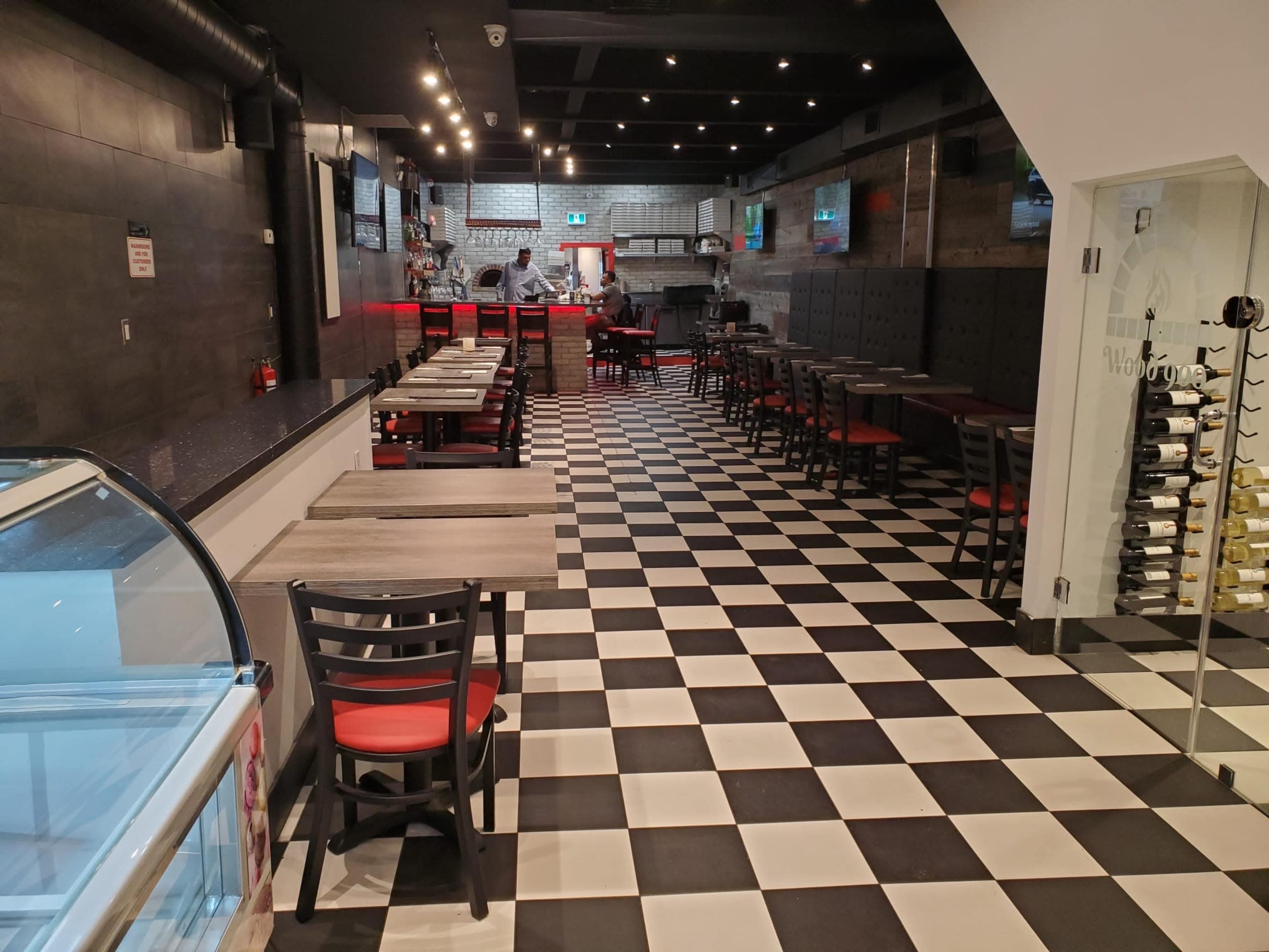 A restaurant interior with black and white checkered flooring, red-accented tables, and a visible kitchen area in the background.