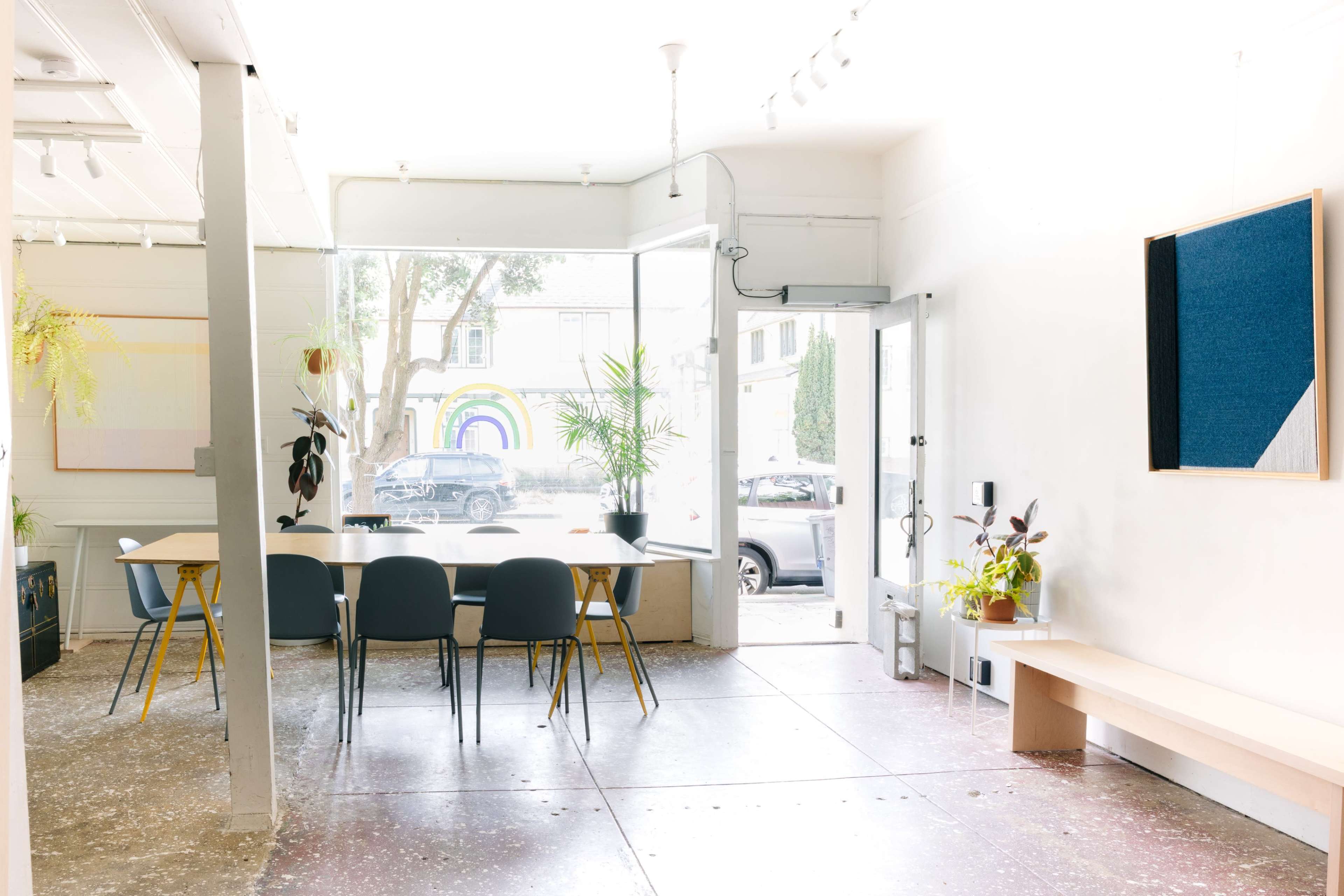 The image shows a bright, minimalist interior of a space featuring a dining table with chairs, large windows allowing natural light, and a view of a street with greenery outside.