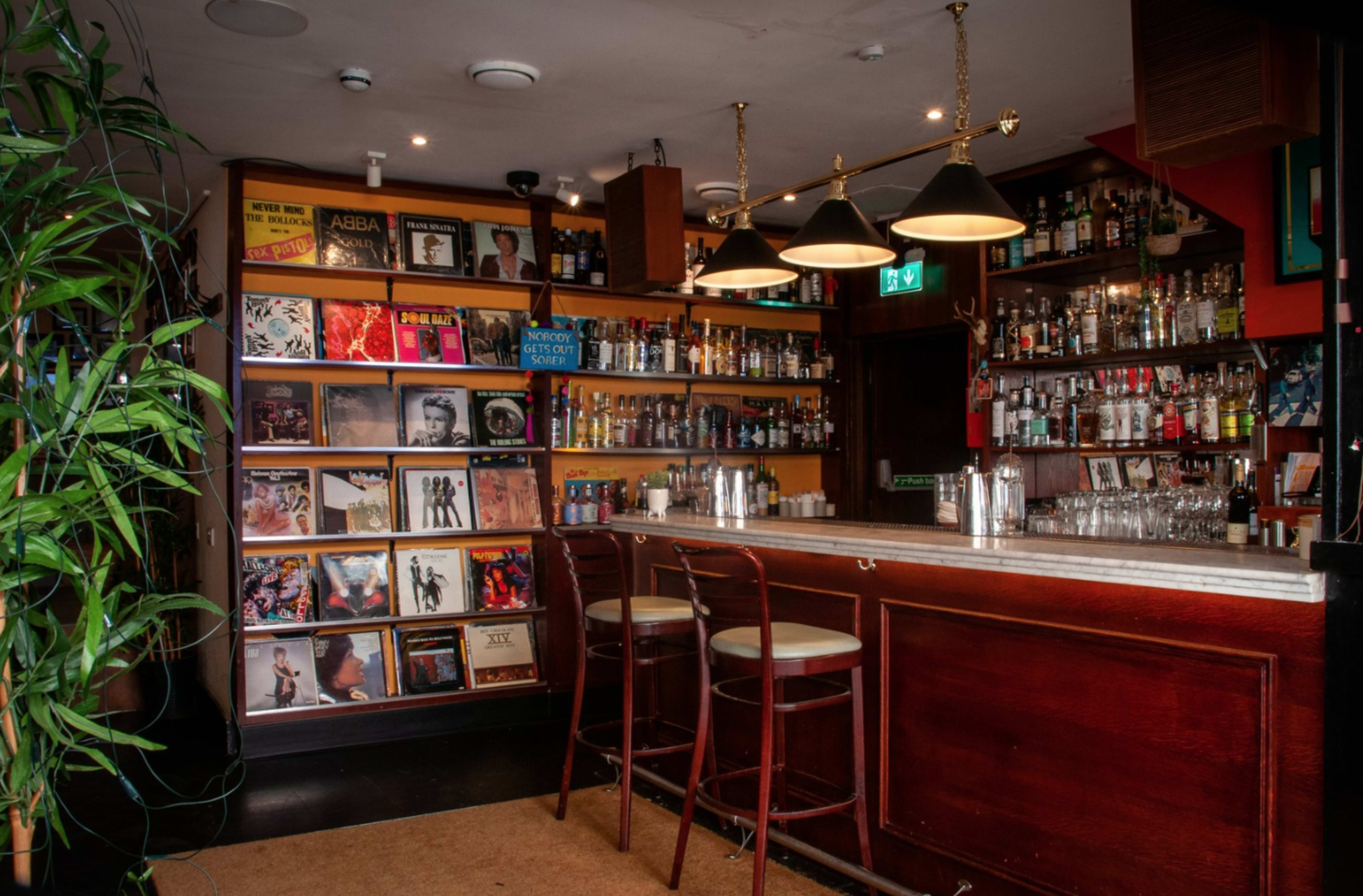 The image depicts a cozy bar interior featuring a polished wooden counter, a wall of vinyl records, and shelves stocked with various bottles of alcohol under warm lighting.
