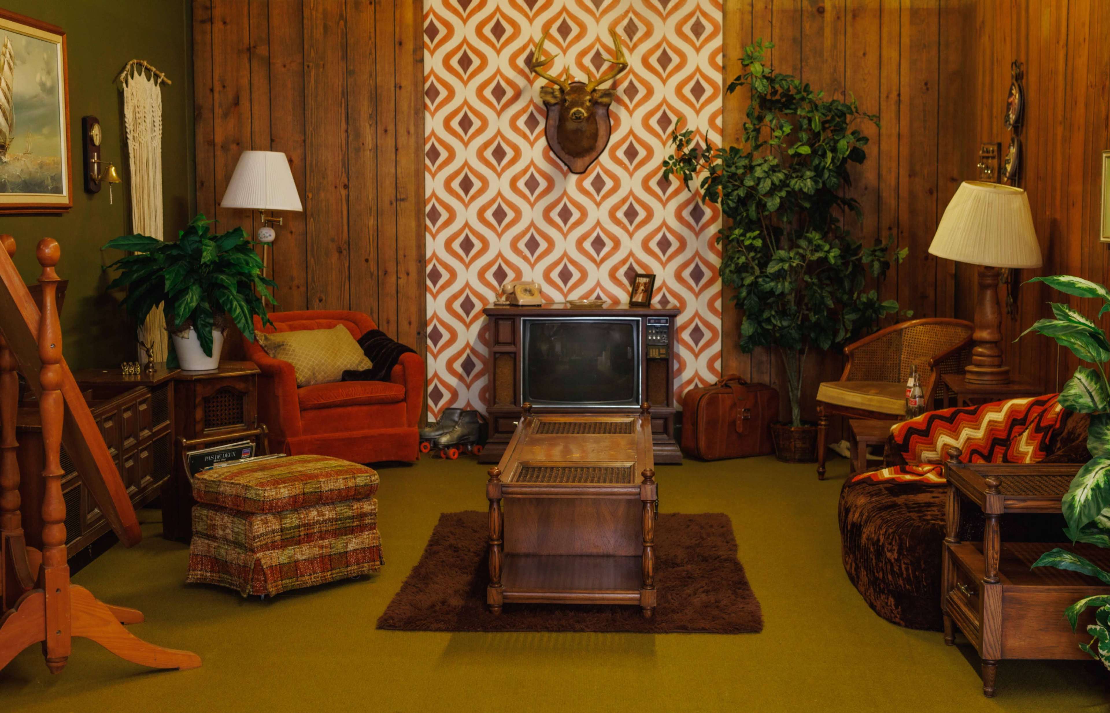The image depicts a retro living room with wooden paneling, patterned wallpaper, and vintage furniture including an orange sofa, a wooden coffee table, and a television.