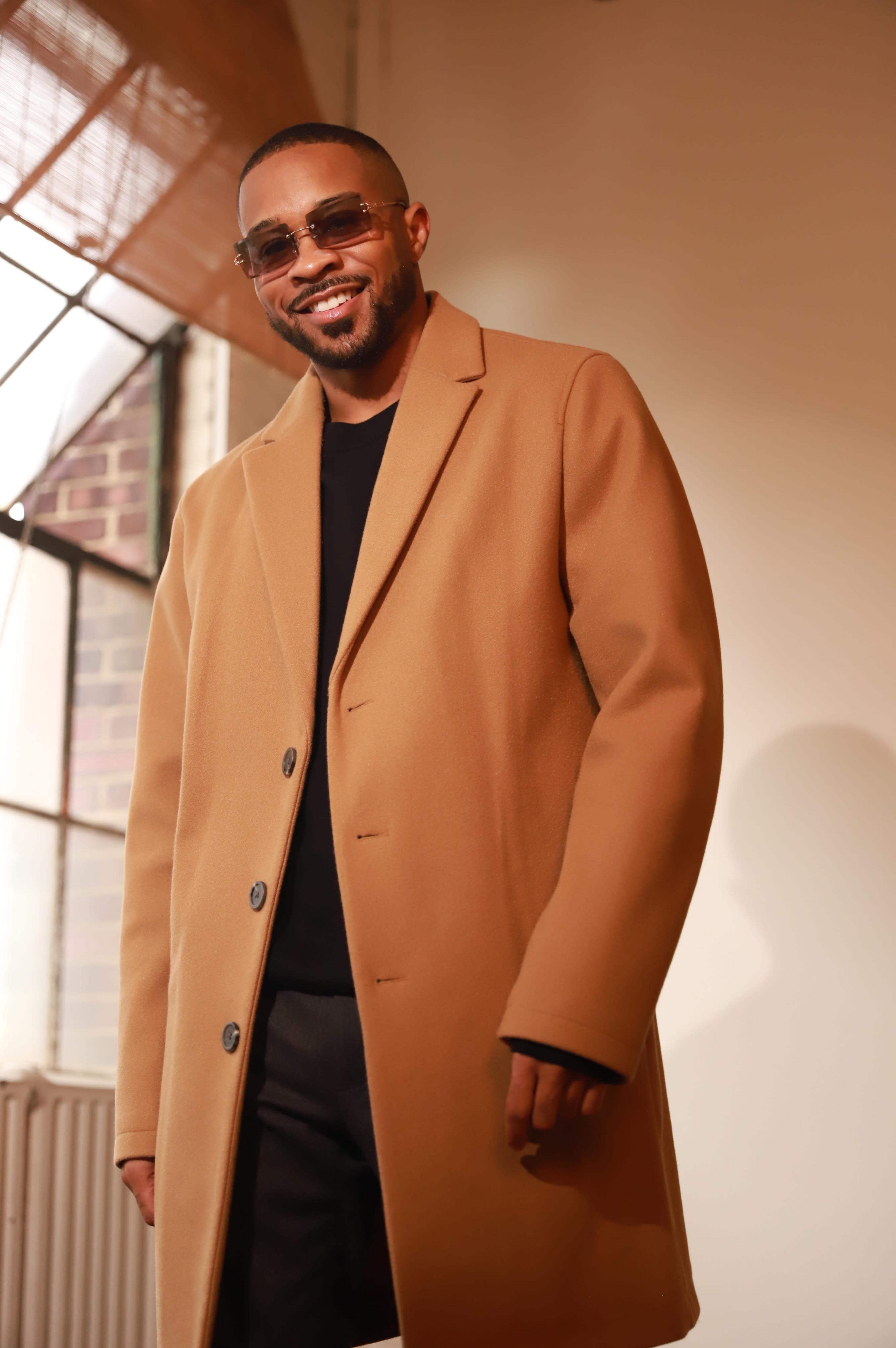 A man wearing a tan coat and sunglasses smiles in front of a large window with natural light streaming in.