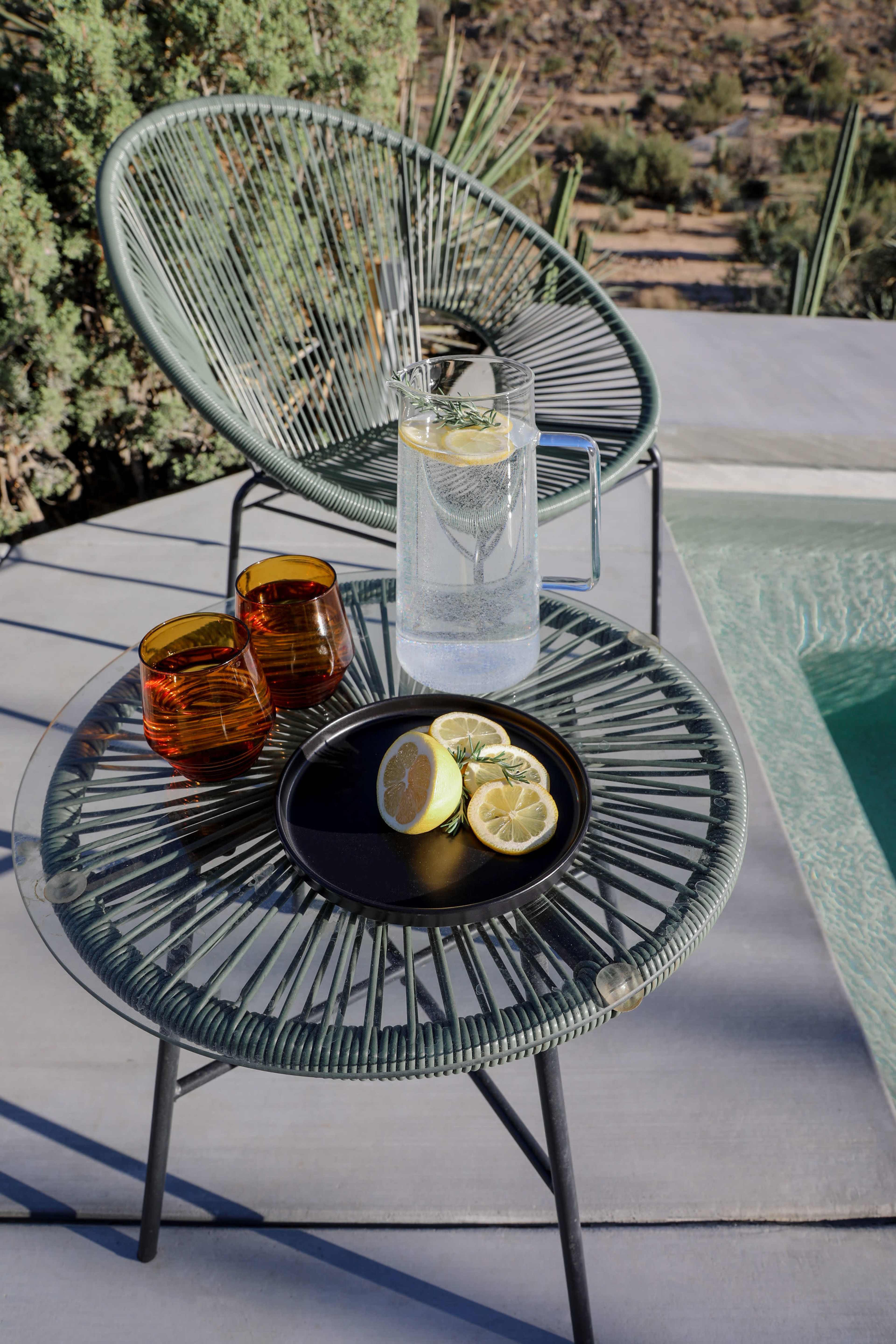 Two chairs with a glass of water and lemon slices on a tray sit beside a pool in a desert landscape.