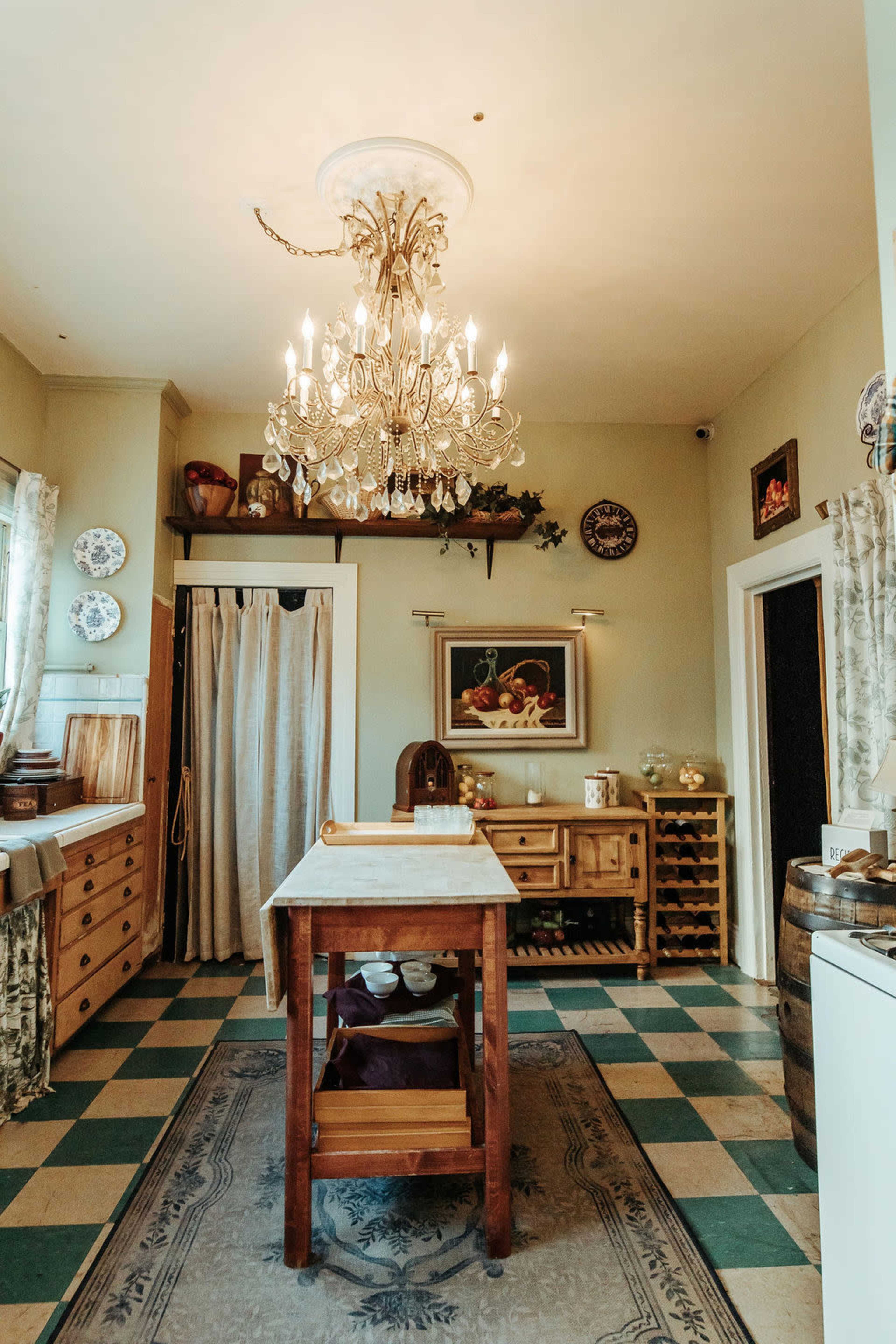 The image shows a kitchen with a wooden table in the center, surrounded by cabinets, a chandelier hanging from the ceiling, and various decorative items on the walls.