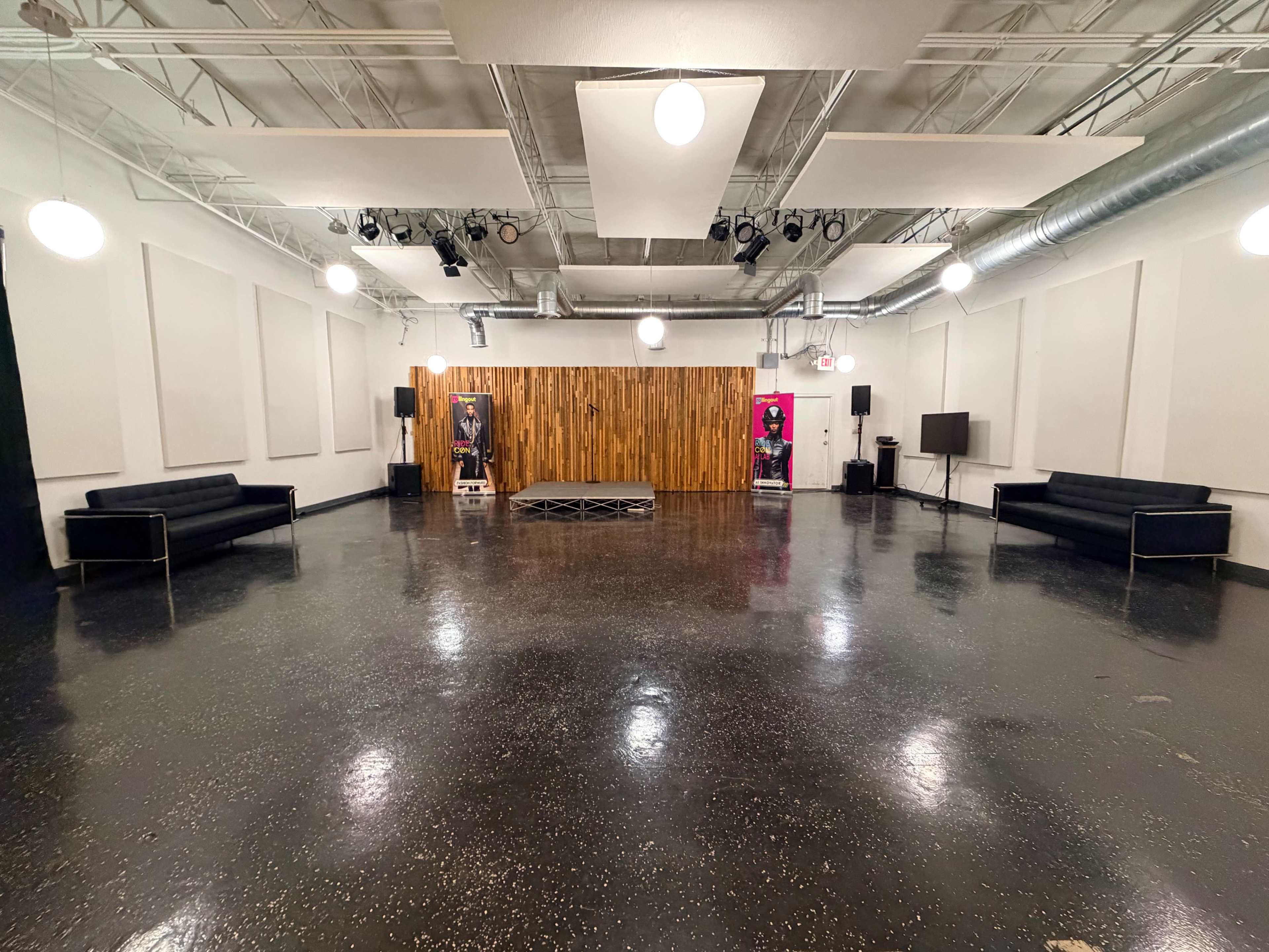 The image shows a spacious room with a polished floor, featuring black sofas along the sides, a wooden wall at the back, and overhead lighting.