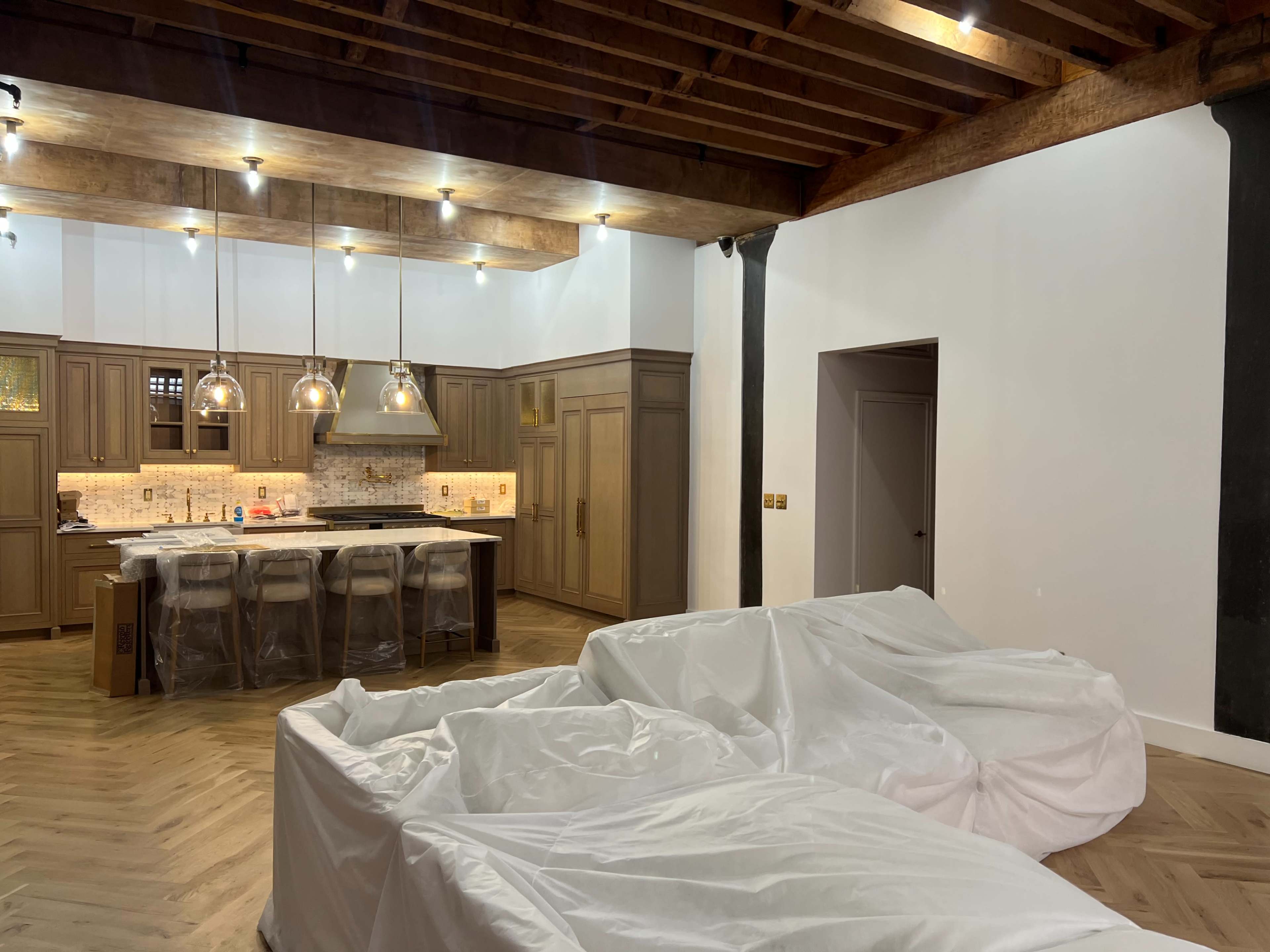 The image shows a partially furnished room featuring two large, white, covered furniture pieces, a kitchen area with wooden cabinetry and pendant lighting, and exposed wooden beams on the ceiling.
