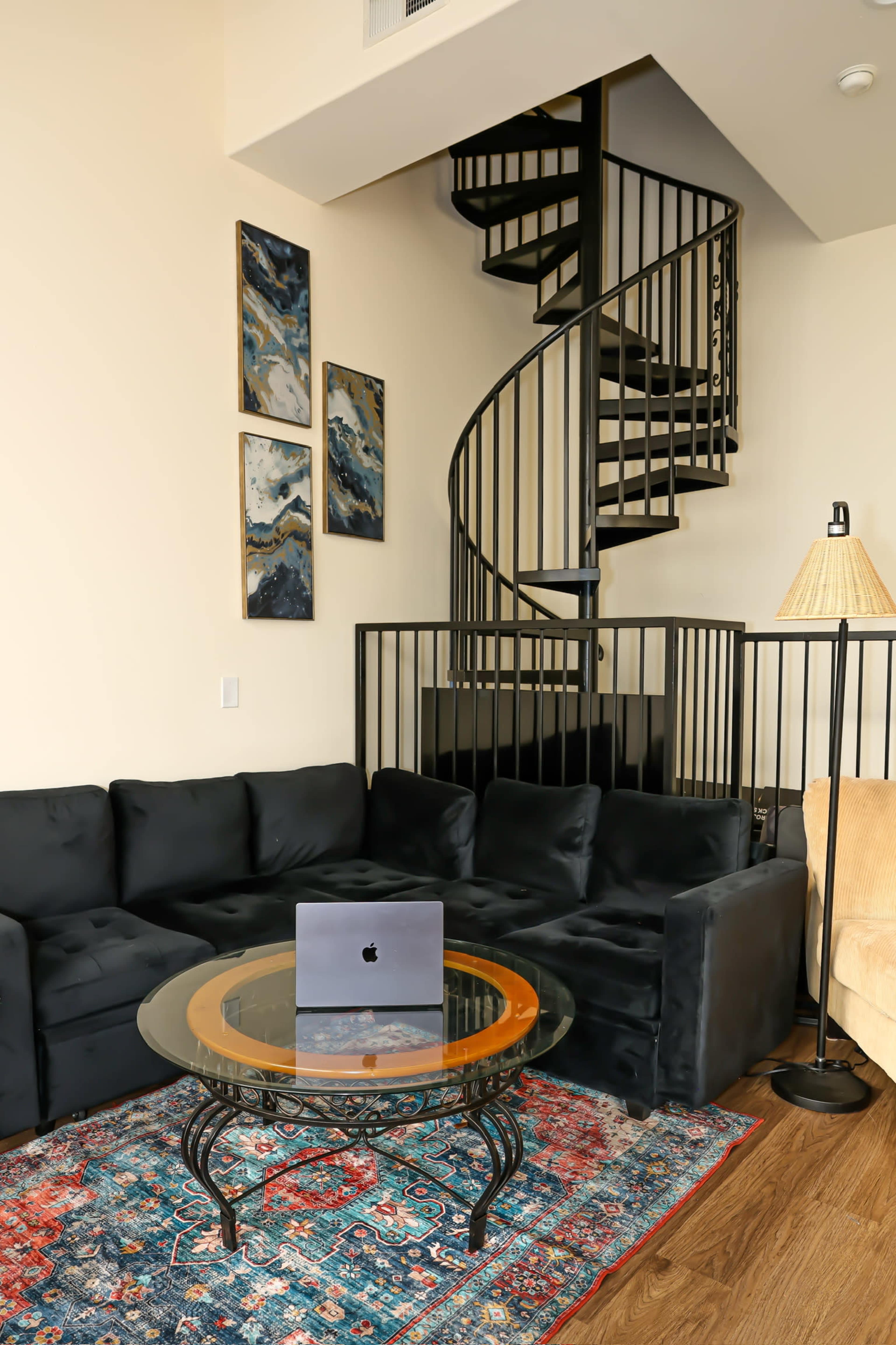 The image shows a modern living room featuring a black sectional couch, a glass coffee table with a laptop on it, a colorful area rug, and a spiral staircase in the background.