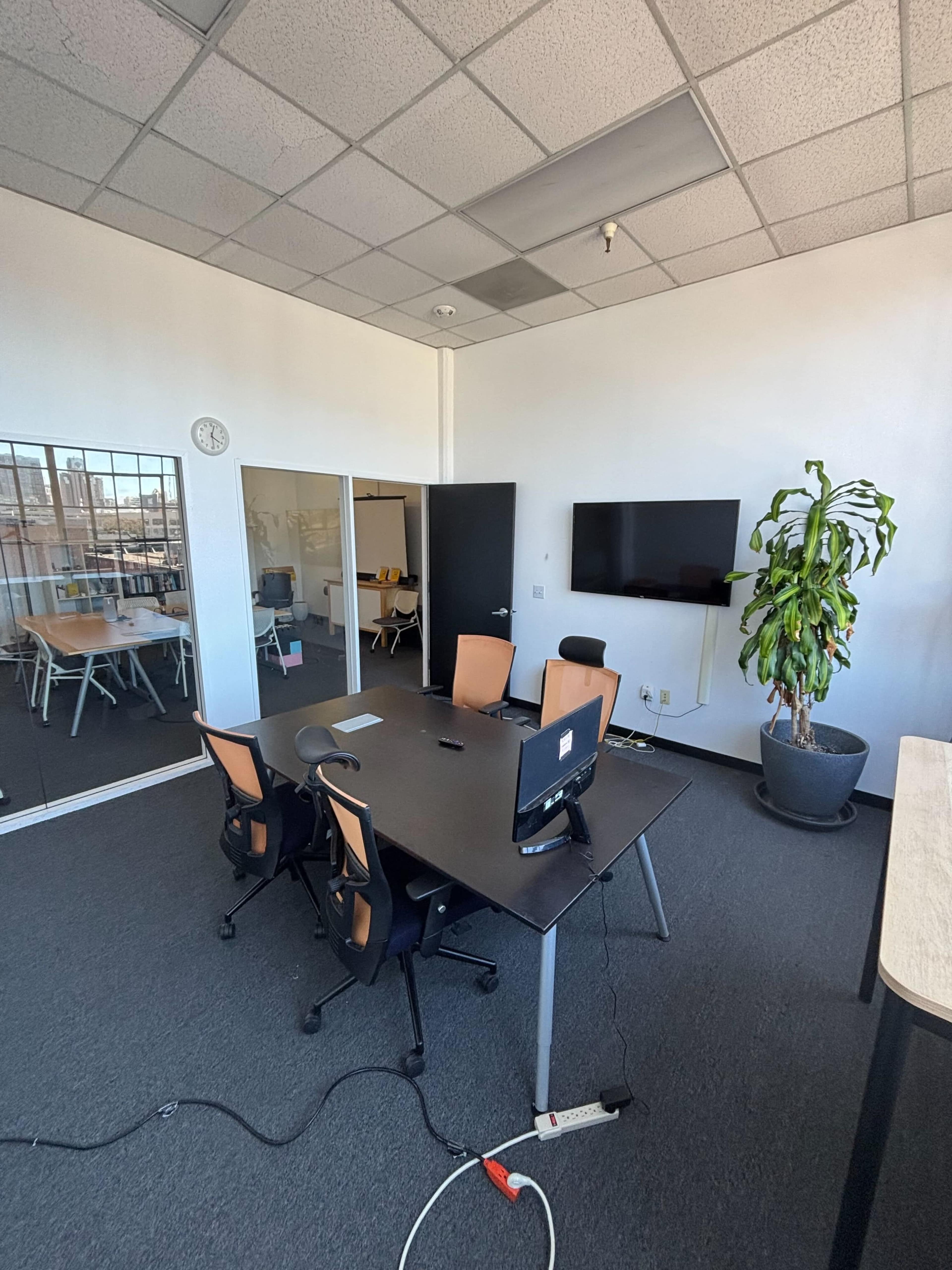 The image shows a modern office space featuring a conference table with four chairs, a television mounted on the wall, and a green plant in a pot.