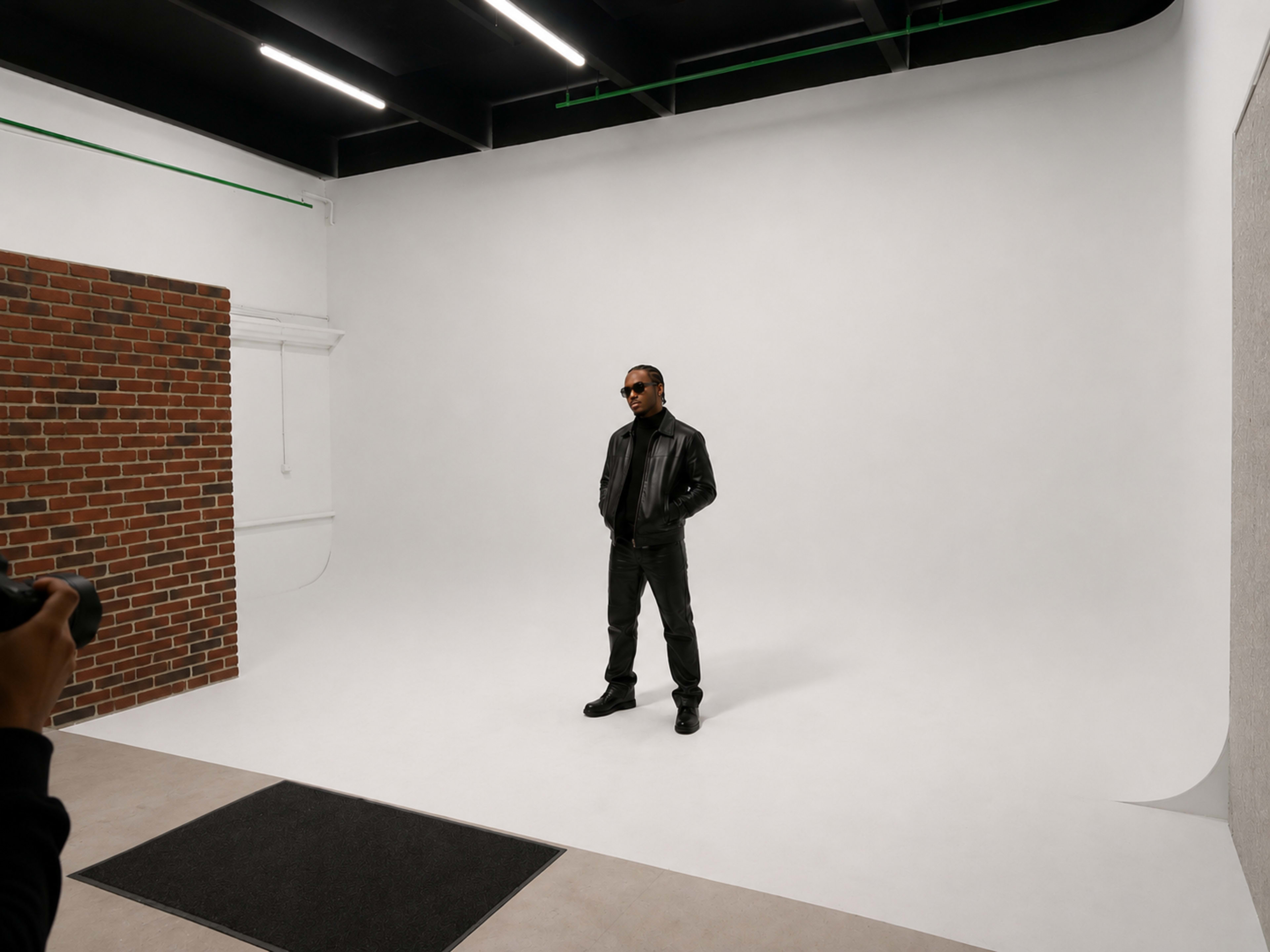 A person stands confidently in a photo studio with a plain white backdrop and a brick wall on one side.