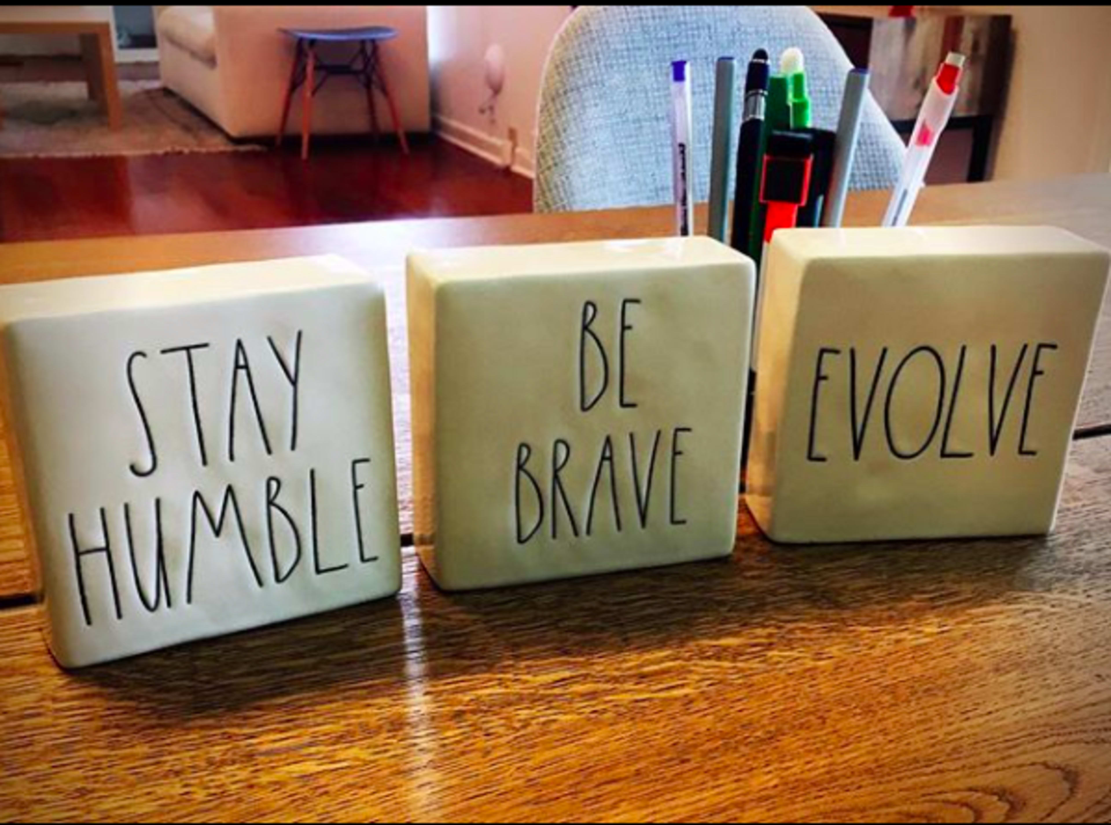 Three decorative tiles on a wooden table, each inscribed with motivational phrases: "STAY HUMBLE," "BE BRAVE," and "EVOLVE."