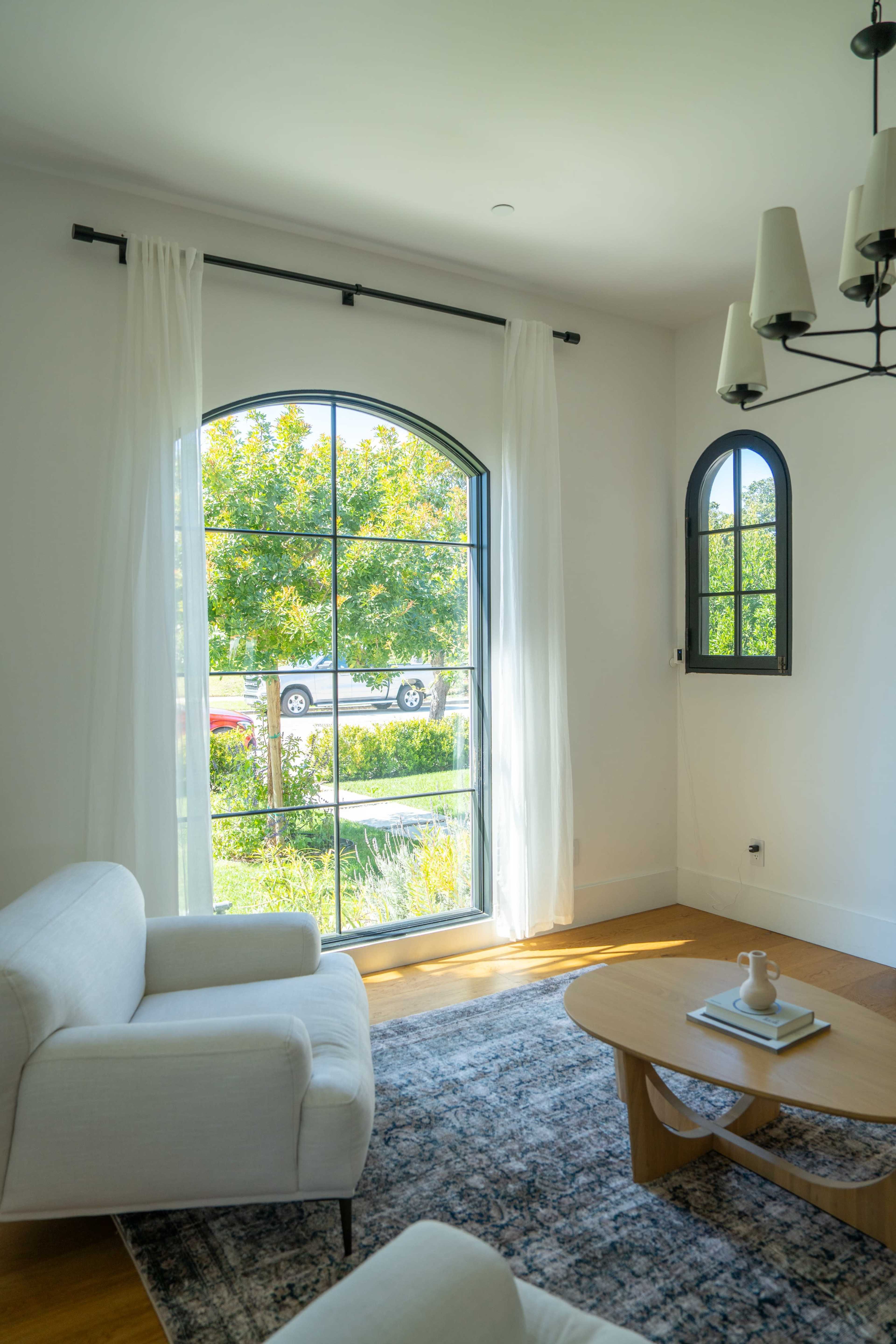 A bright room with two white sofas, a wooden coffee table, and large windows that overlook a garden and a street.