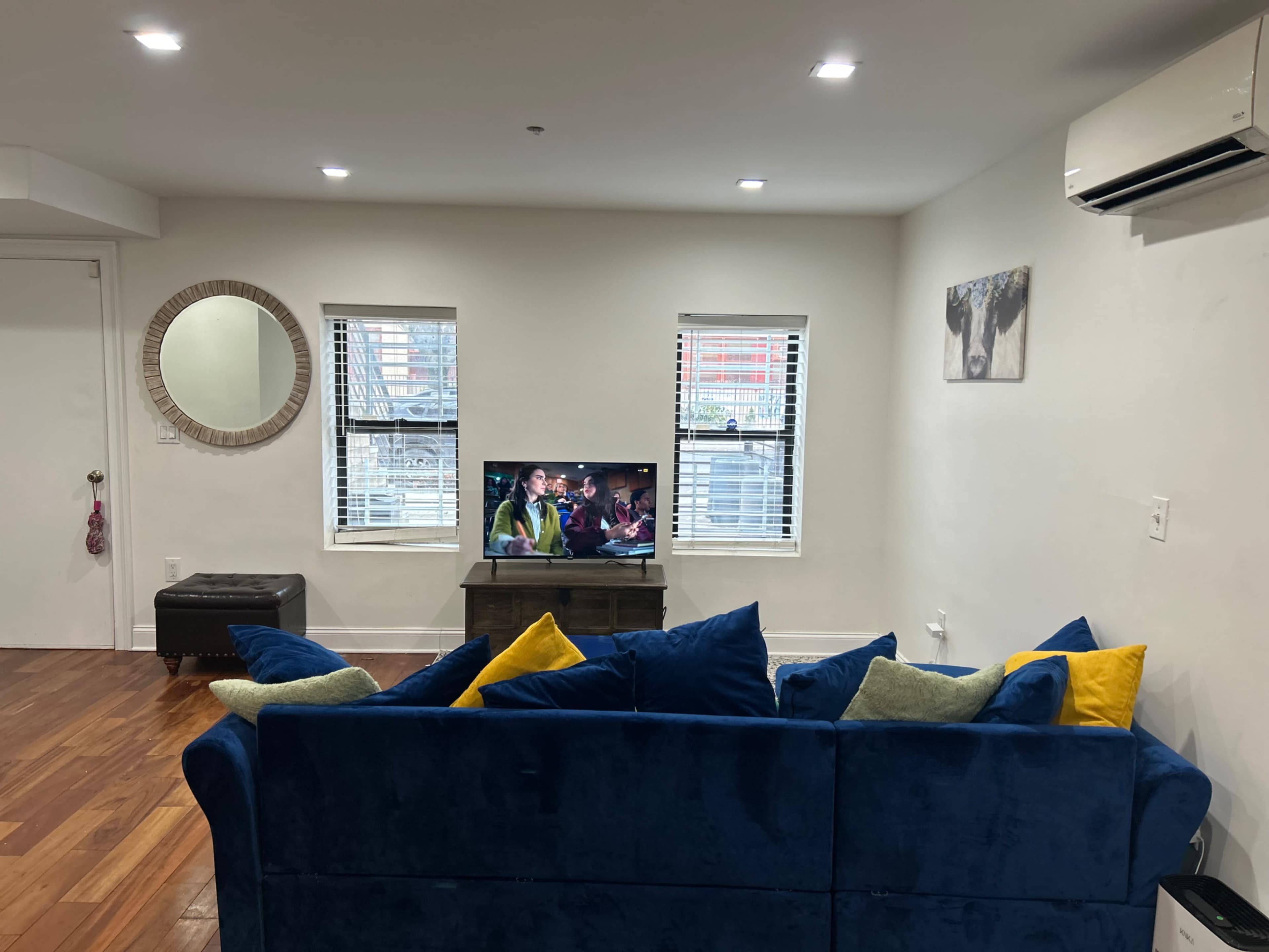 A blue sofa with colorful cushions faces a television in a living room with hardwood floors and two windows.