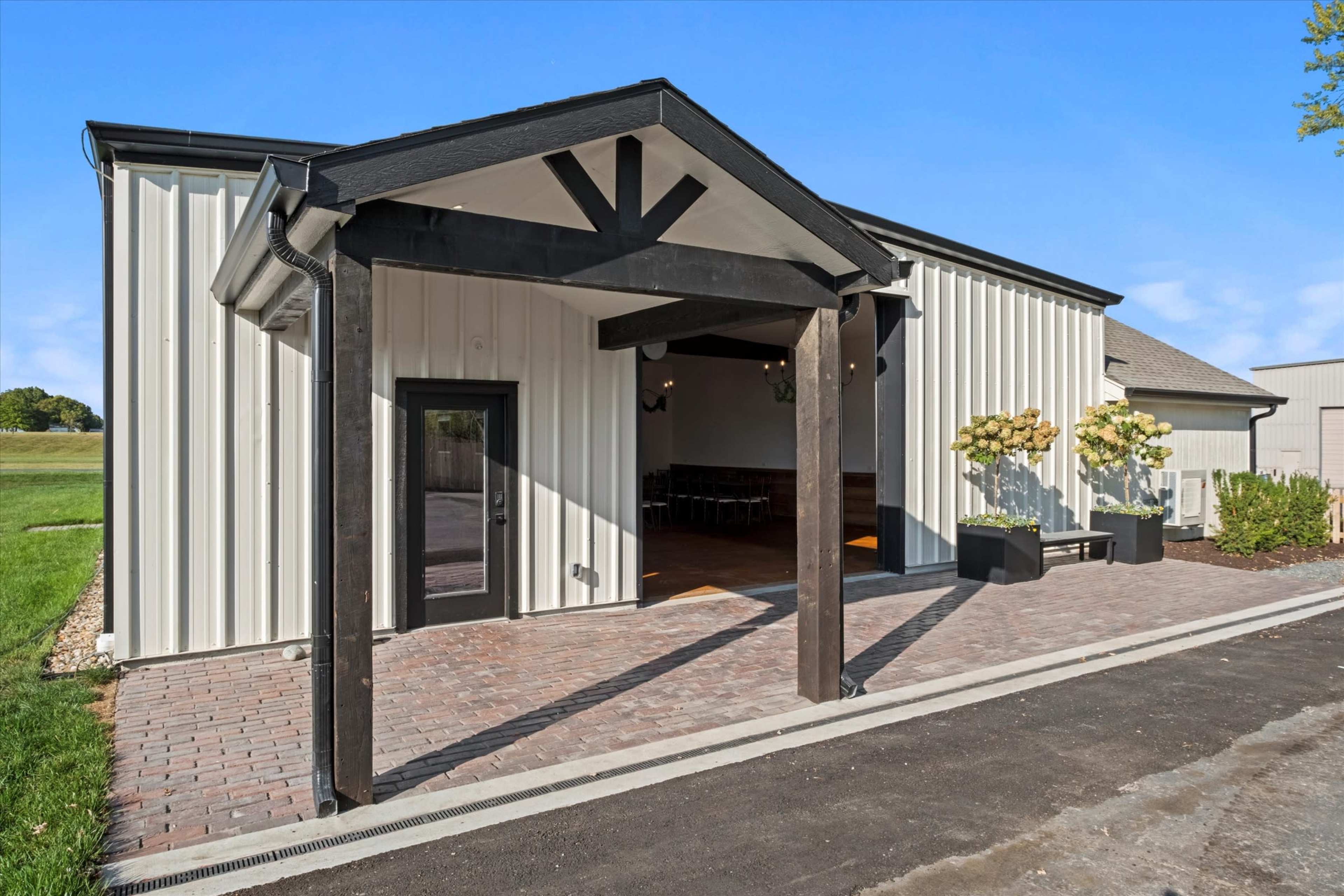The image shows a modern building with a metal exterior, featuring a covered entrance supported by wooden beams and surrounded by landscaped areas.