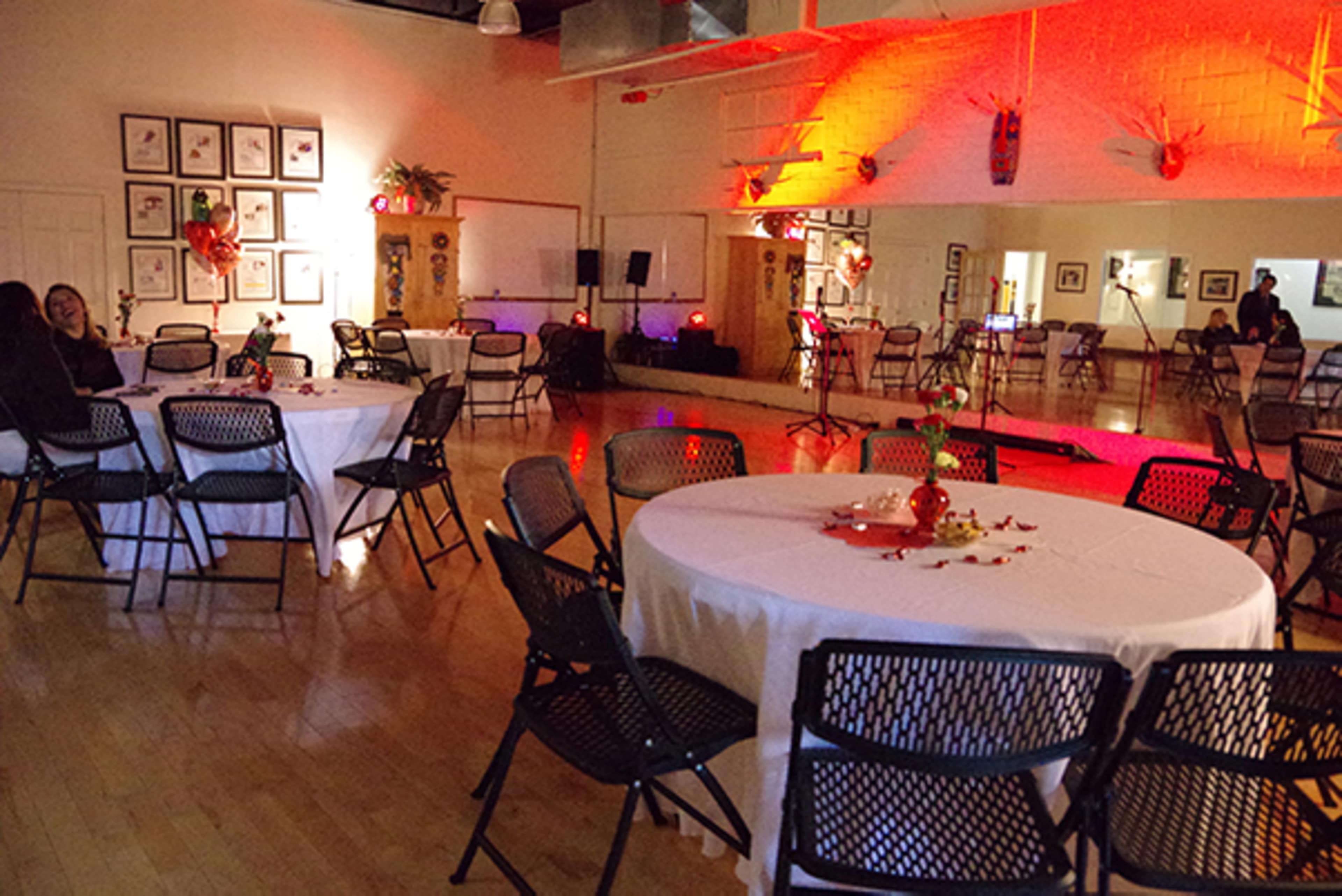 A large indoor space set up for an event, featuring round tables with white tablecloths and chairs arranged around them, with decorations and soft lighting illuminating the area.