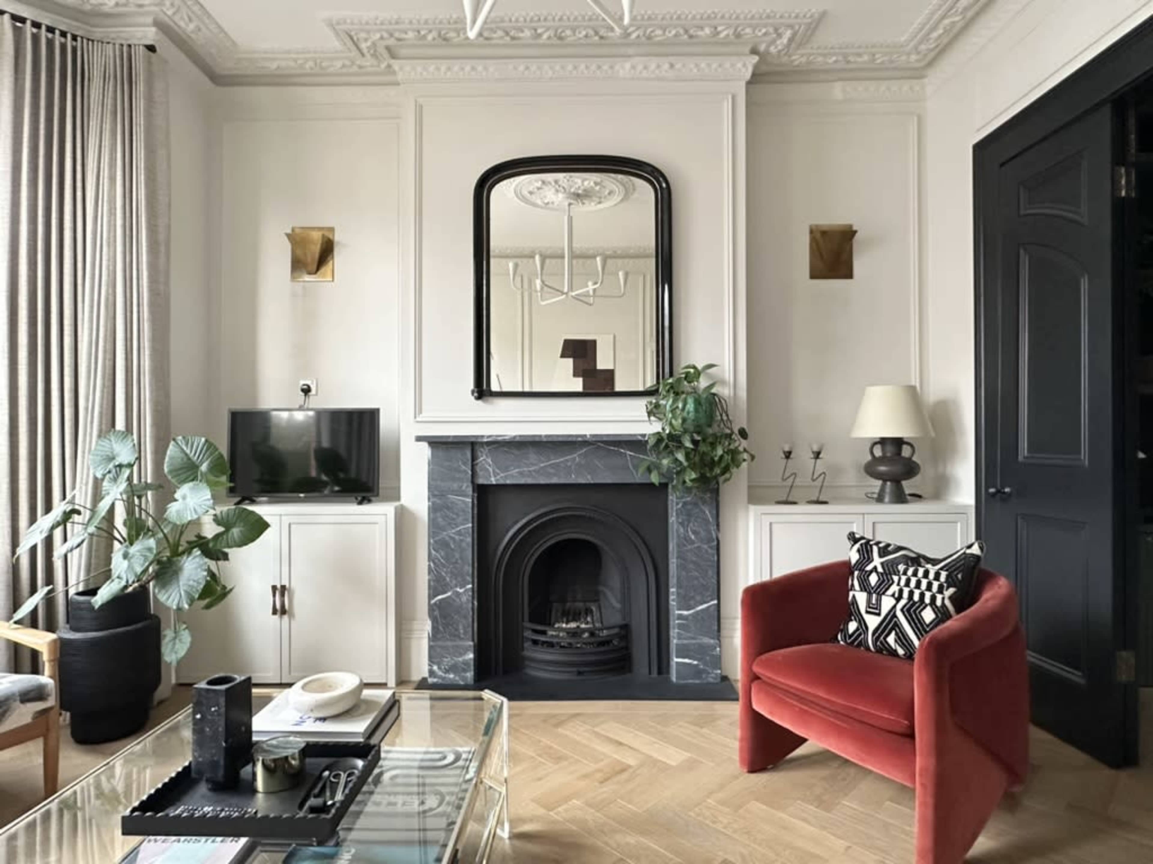 The image shows a stylish living room featuring a black marble fireplace, a large mirror above it, a red armchair, and decorative houseplants.