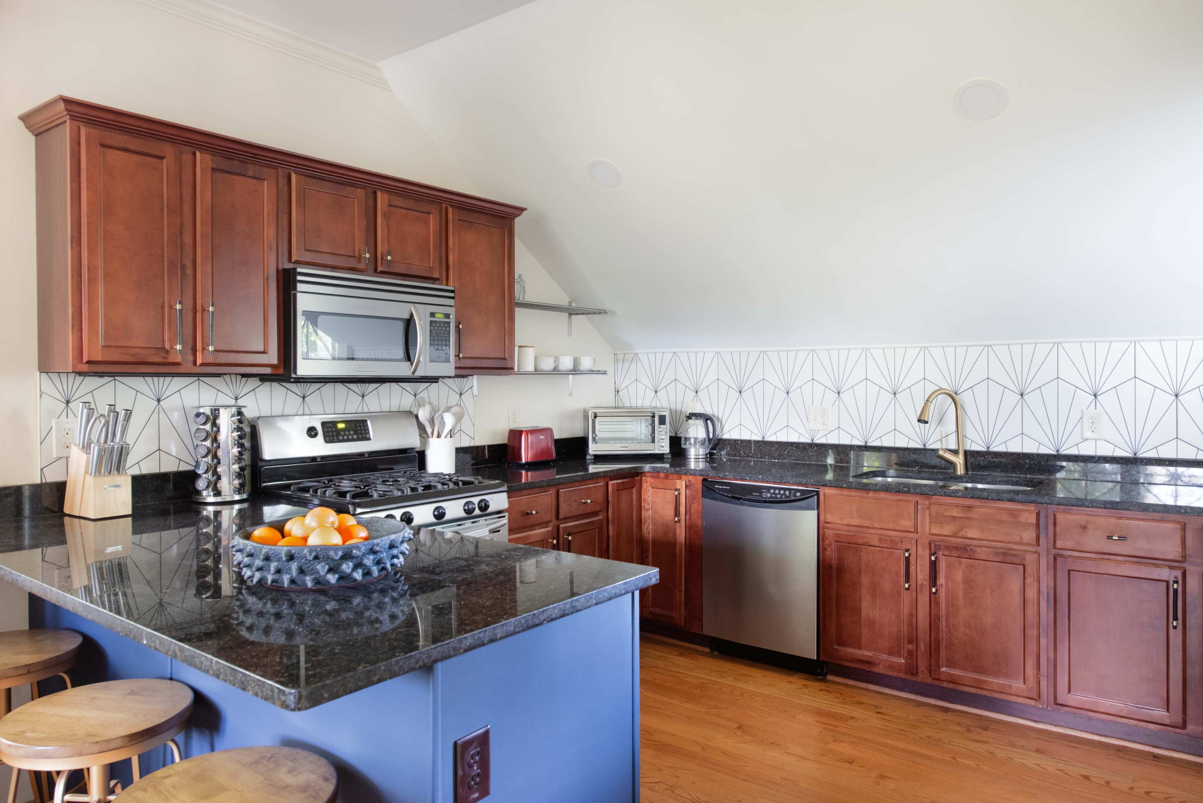 The image shows a modern kitchen featuring dark wood cabinetry, stainless steel appliances, a black granite countertop, and a geometric tile backsplash.
