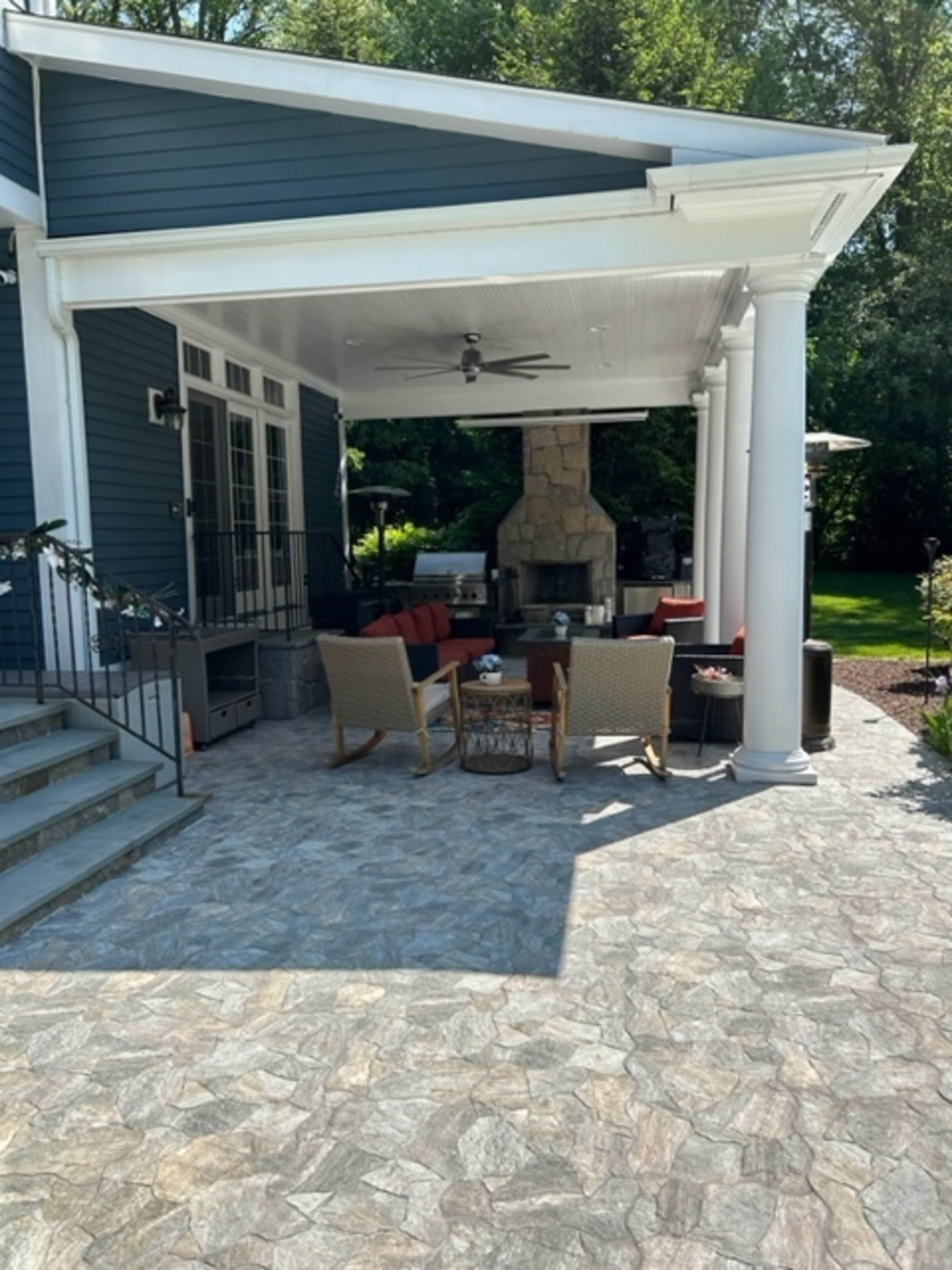 A covered outdoor patio area with two chairs, a coffee table, and a stone fireplace, surrounded by greenery.