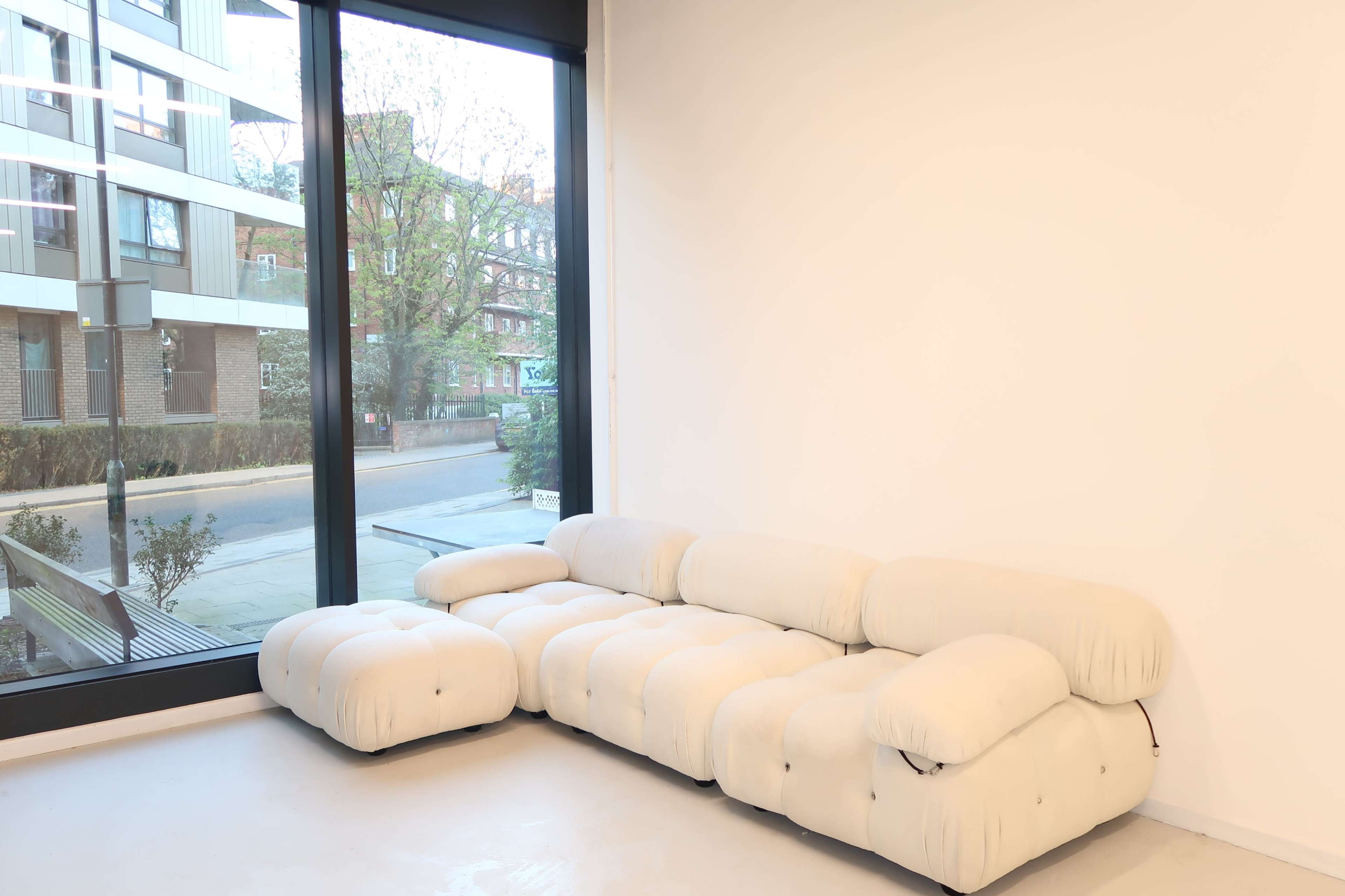 A light-colored, tufted sofa is positioned near a large window overlooking a sidewalk and buildings outside.