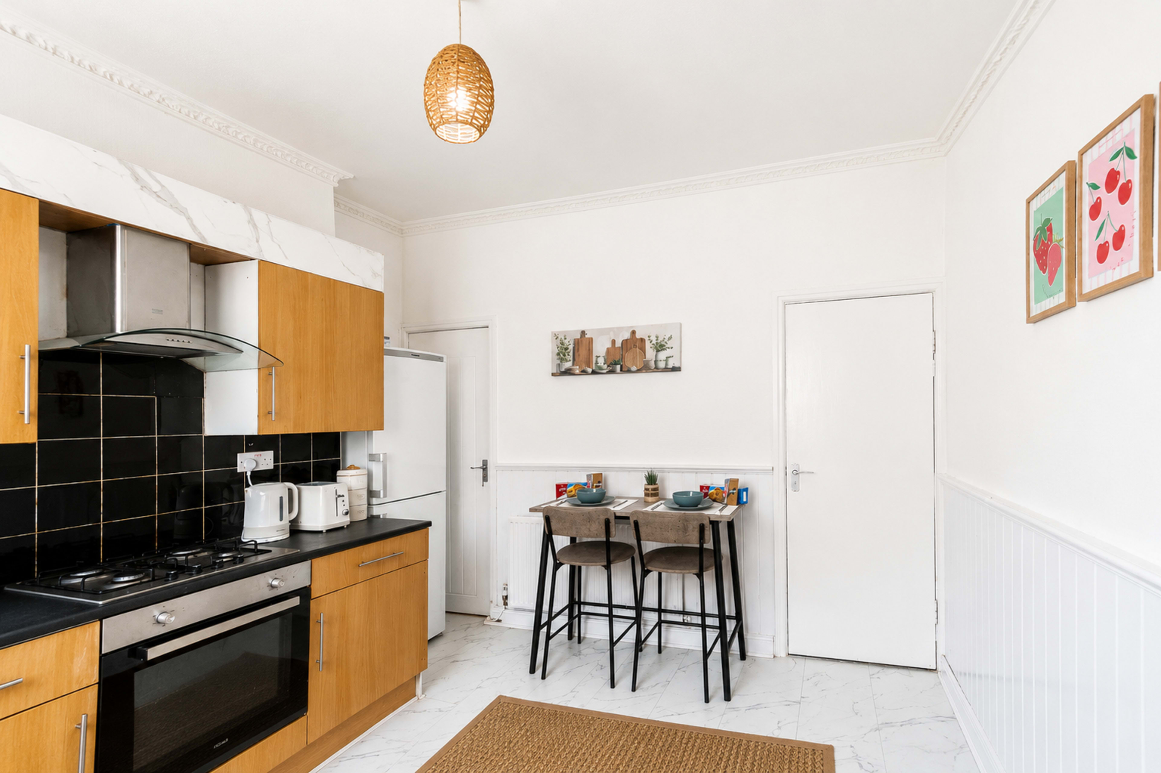 The image shows a modern kitchen featuring wooden cabinets, a black countertop, and a small dining area with two stools.