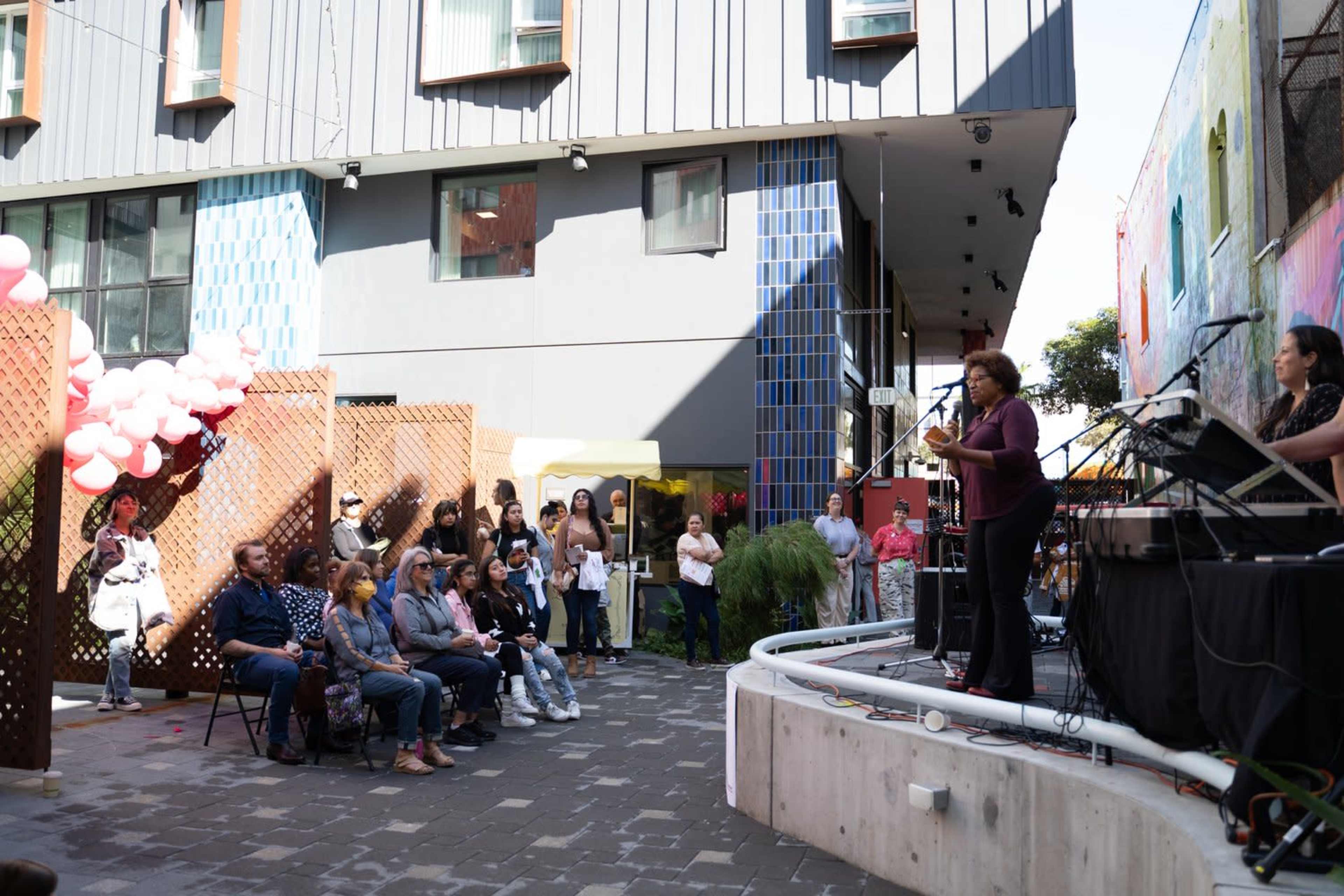 A speaker addresses an audience in an outdoor courtyard with colorful murals and decorations.