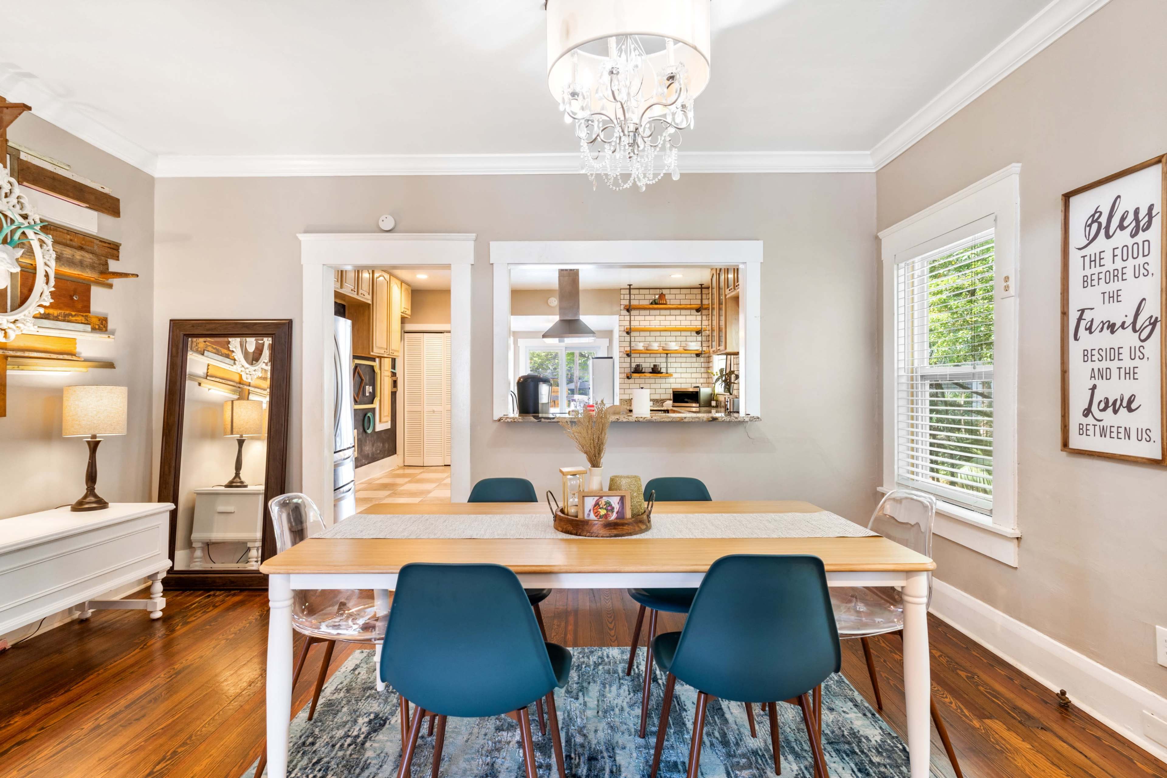A dining area features a wooden table surrounded by blue chairs, with a chandelier overhead and a light-colored wall adorned with decorative elements.