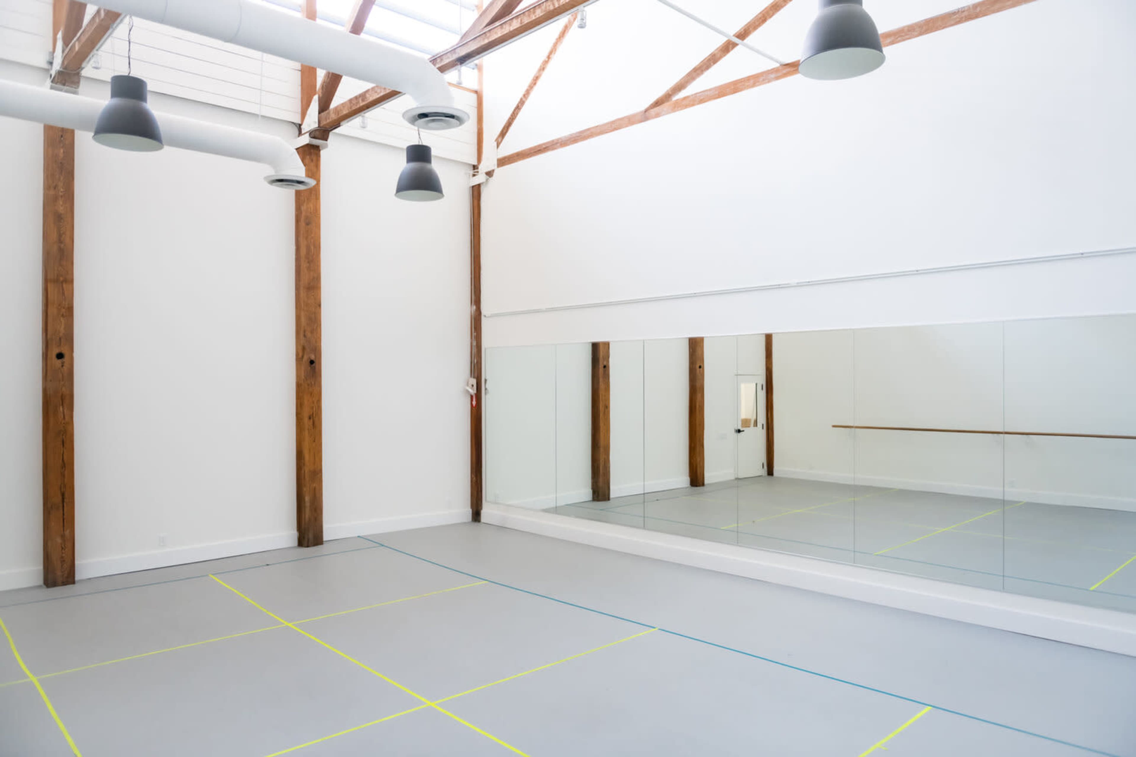 A spacious dance studio features high wooden beams, a mirrored wall, and a light gray floor marked with colored lines.