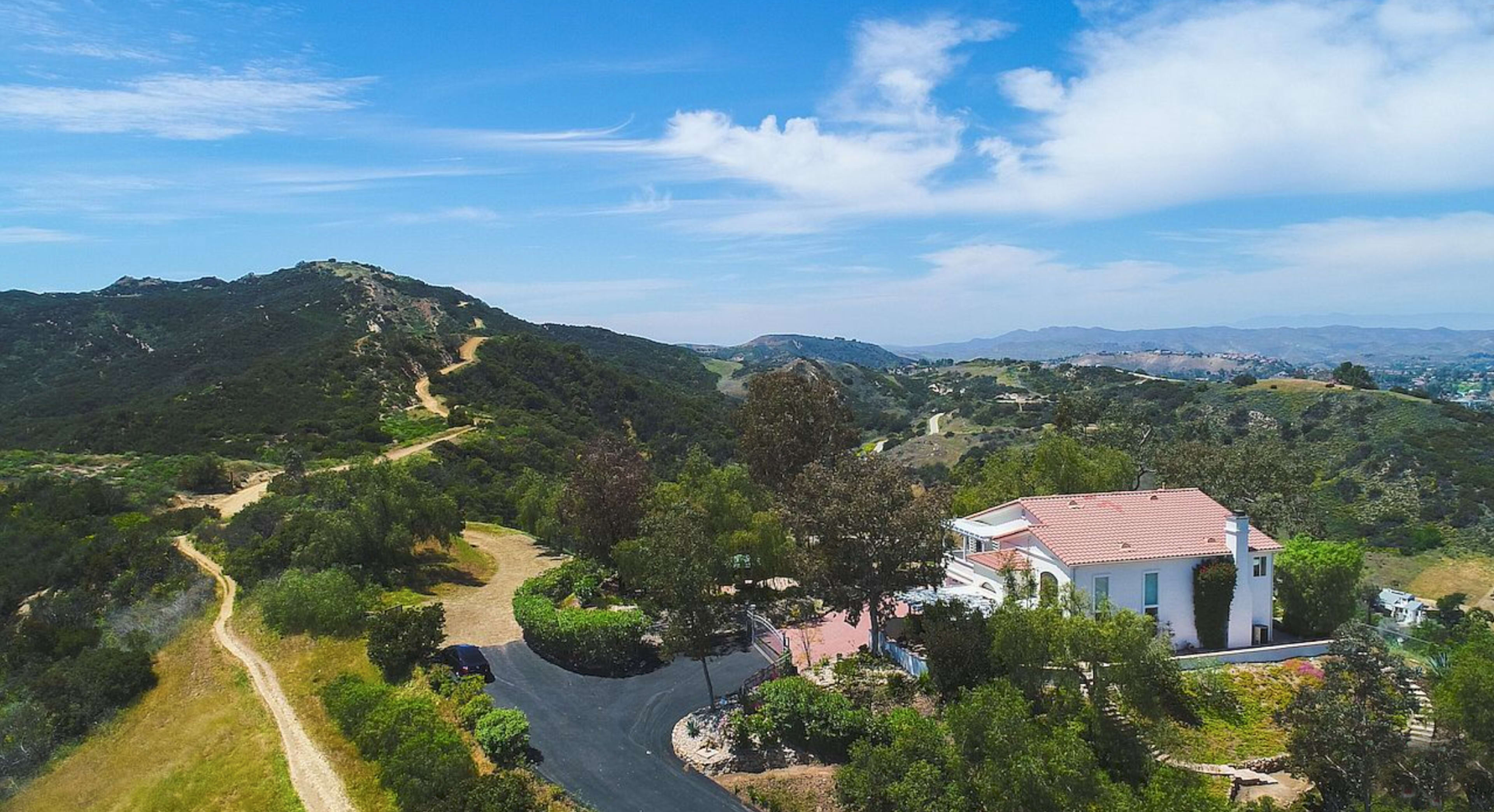 A large white house with a red roof is nestled among green hills, surrounded by a winding driveway and paths leading into the mountains.
