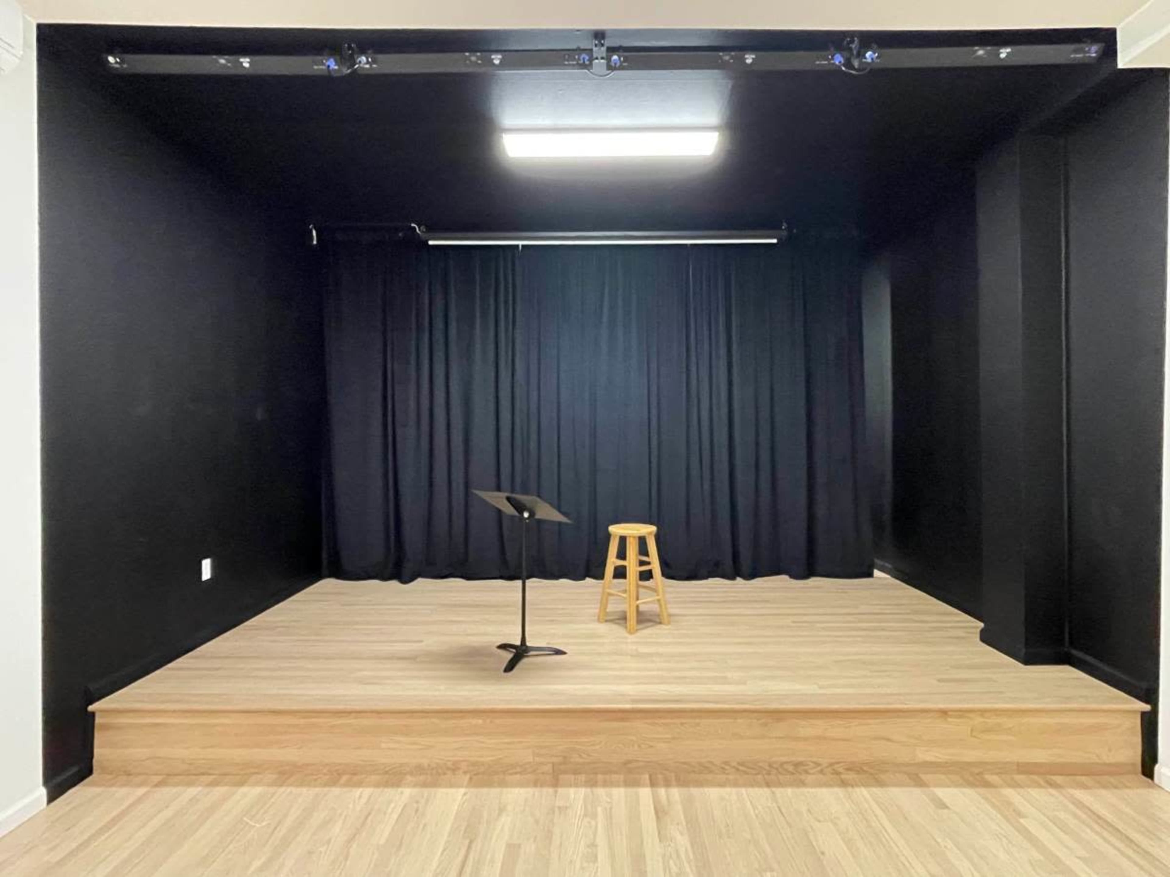 The image shows a small staging area with a wooden platform, a music stand, and a stool, all set against a backdrop of black curtains and black walls.