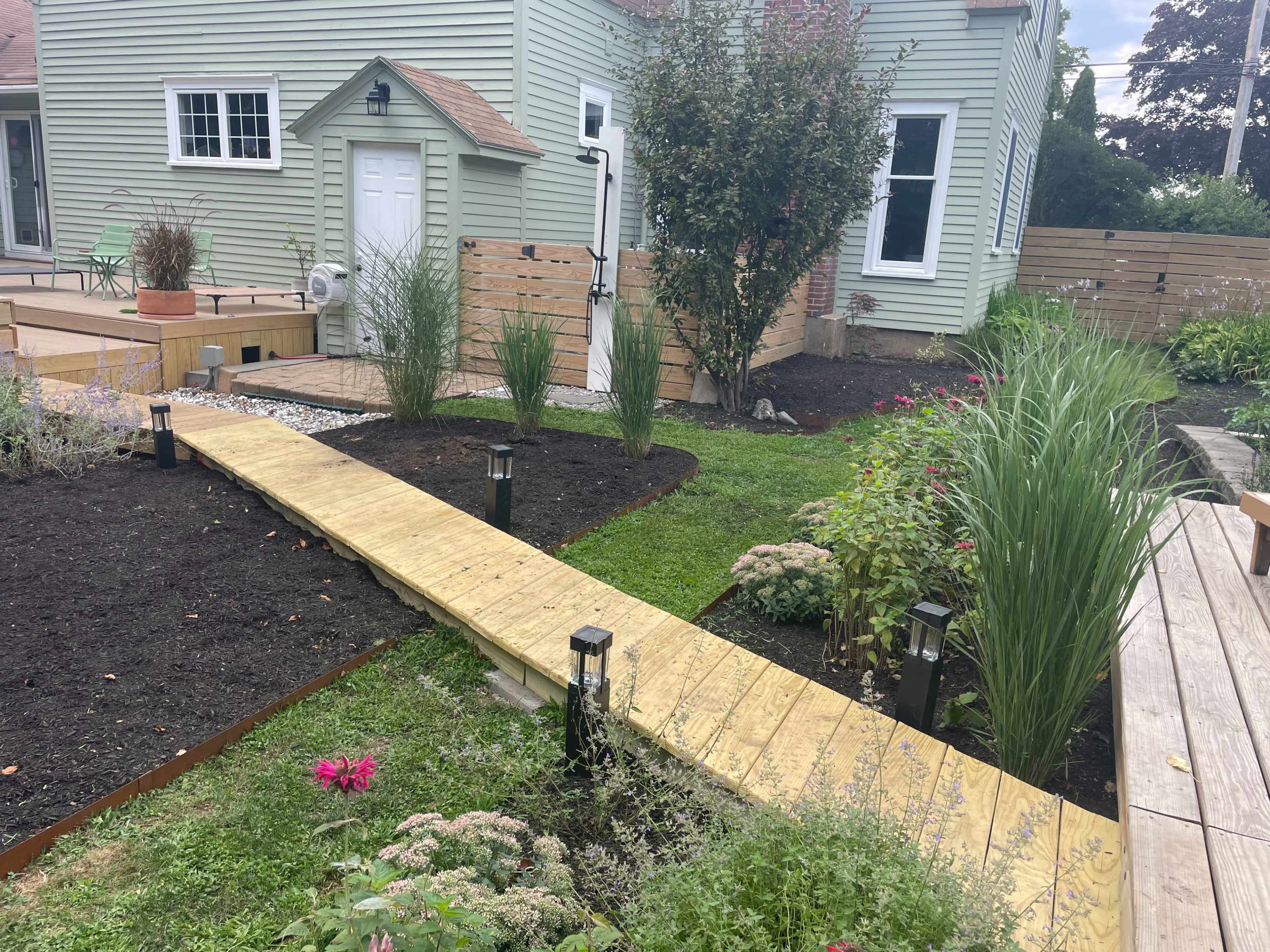 The image shows a landscaped yard featuring a wooden pathway leading to a garden area with various plants, a small building, and decorative lighting along the path.
