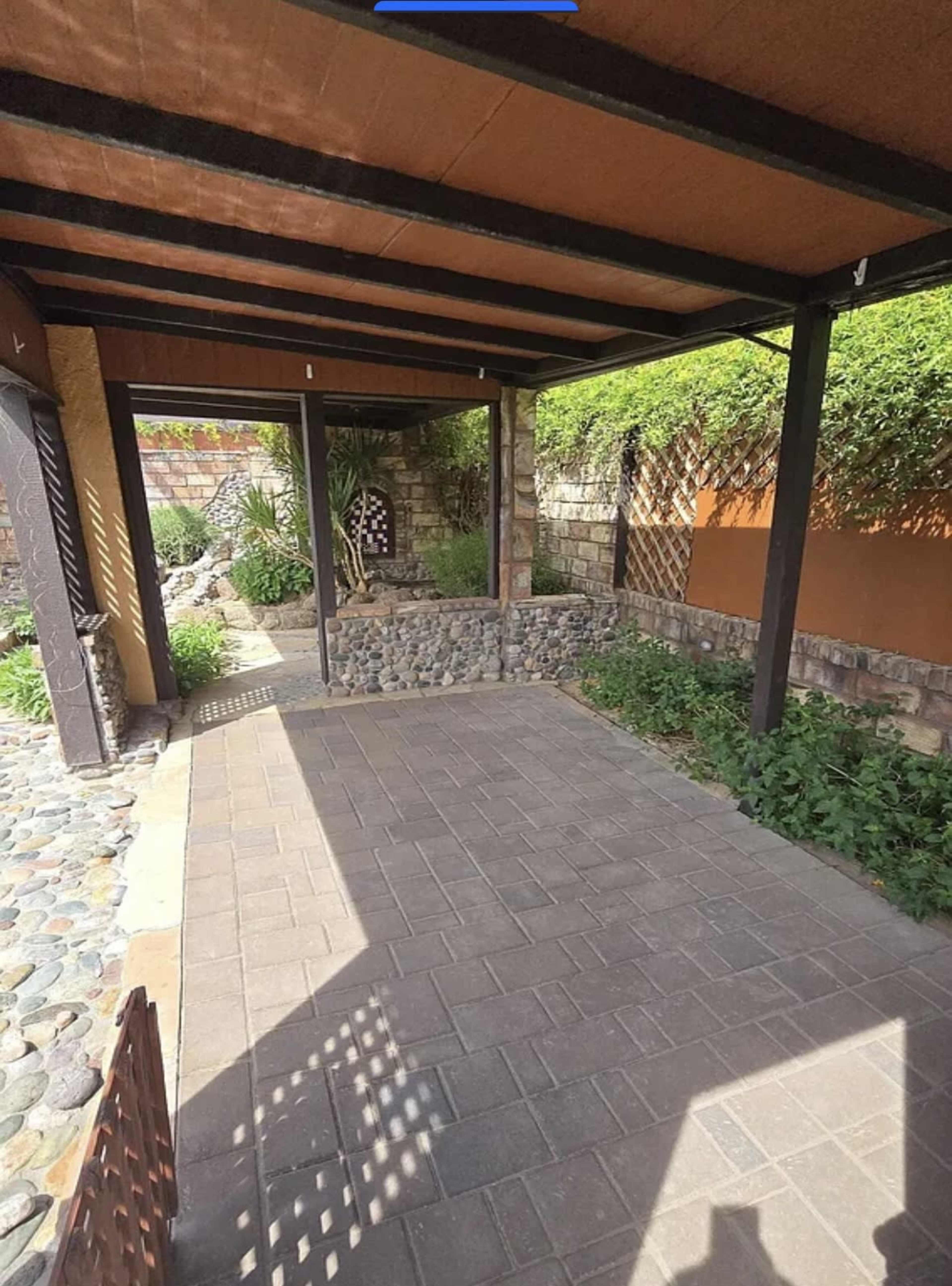 The image shows a covered outdoor patio with stone flooring and a landscaped garden surrounding it.