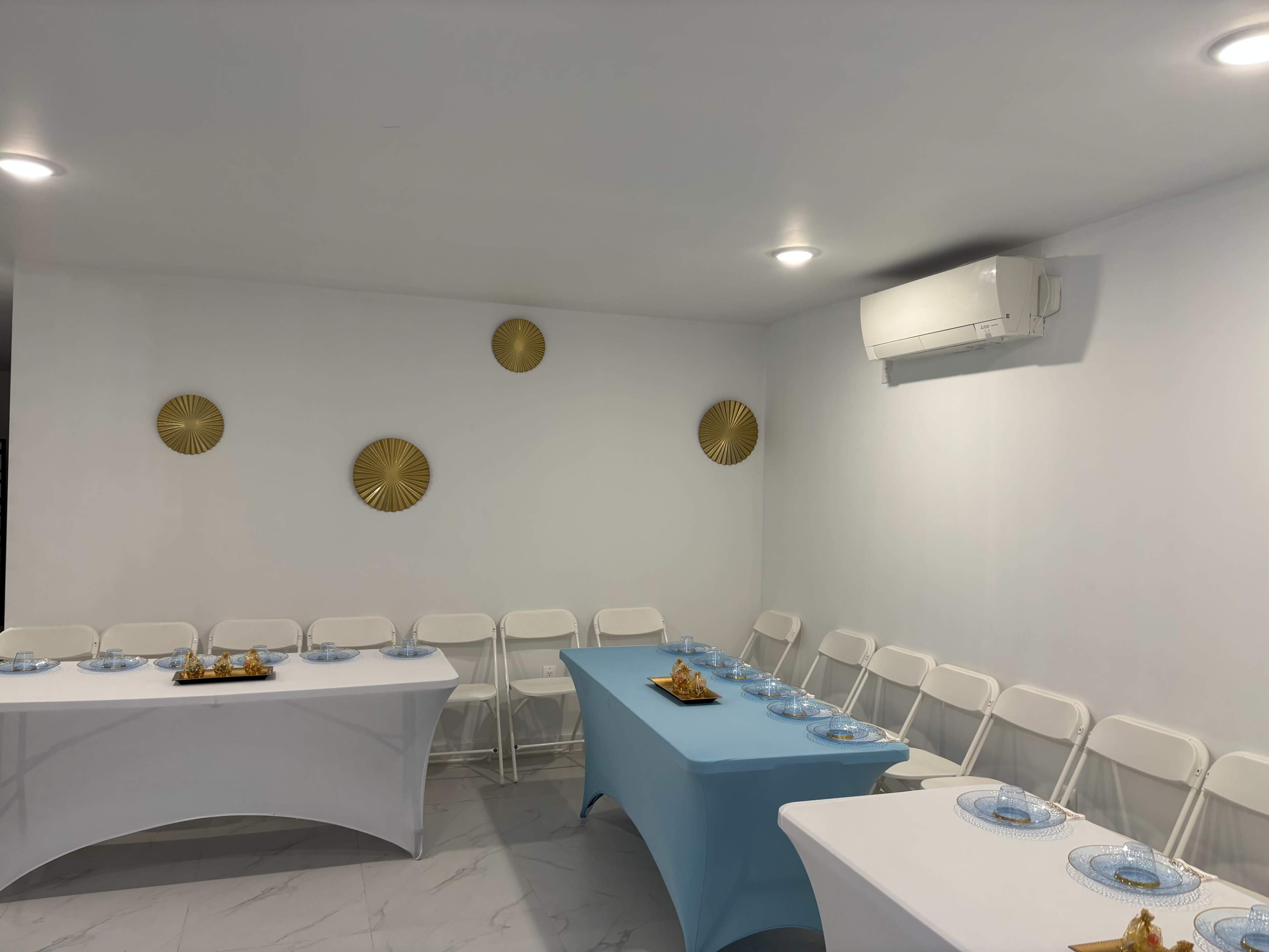 The image shows a small indoor event space with white walls, a light-colored floor, and seating arranged around a blue and white table set with plates.