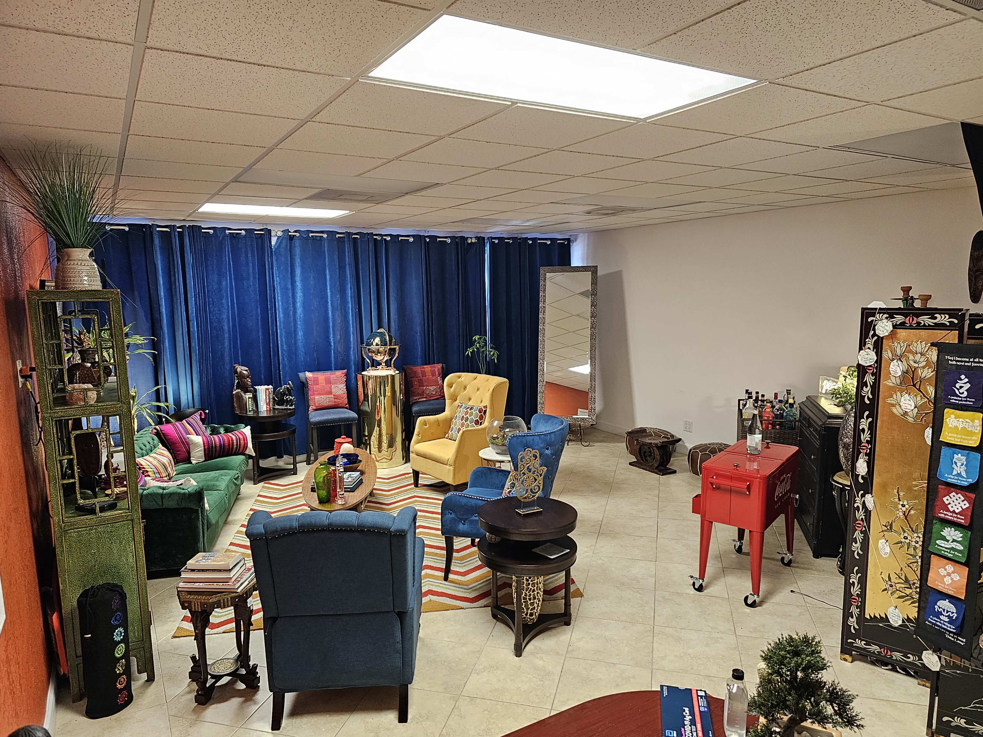 A vibrant room features colorful furniture, including a yellow and blue chair, a striped rug, and a bar cart, with large blue curtains covering a window.