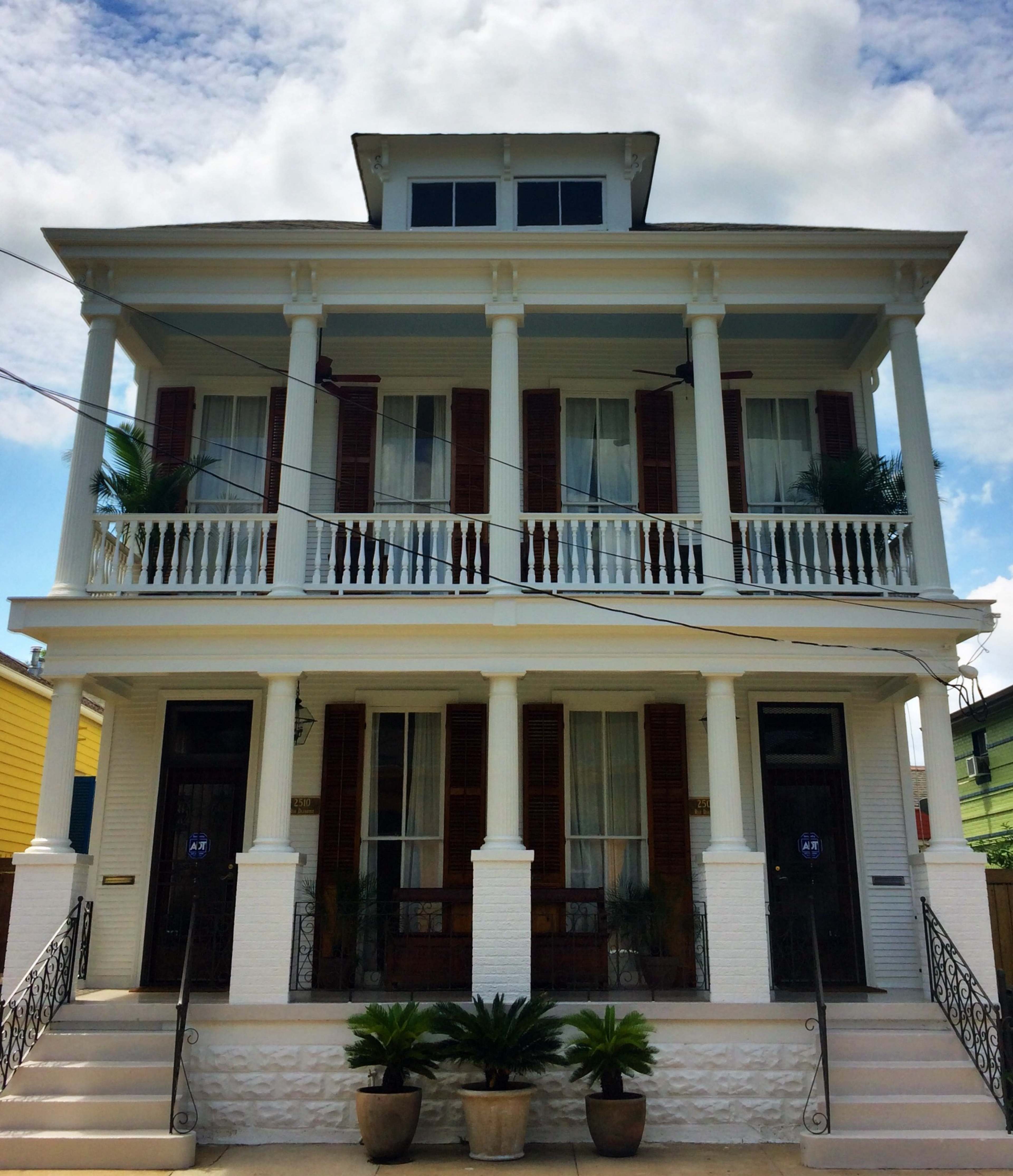 The image shows a two-story white house with a porch supported by six columns, featuring wooden shutters and potted plants at the entrance.