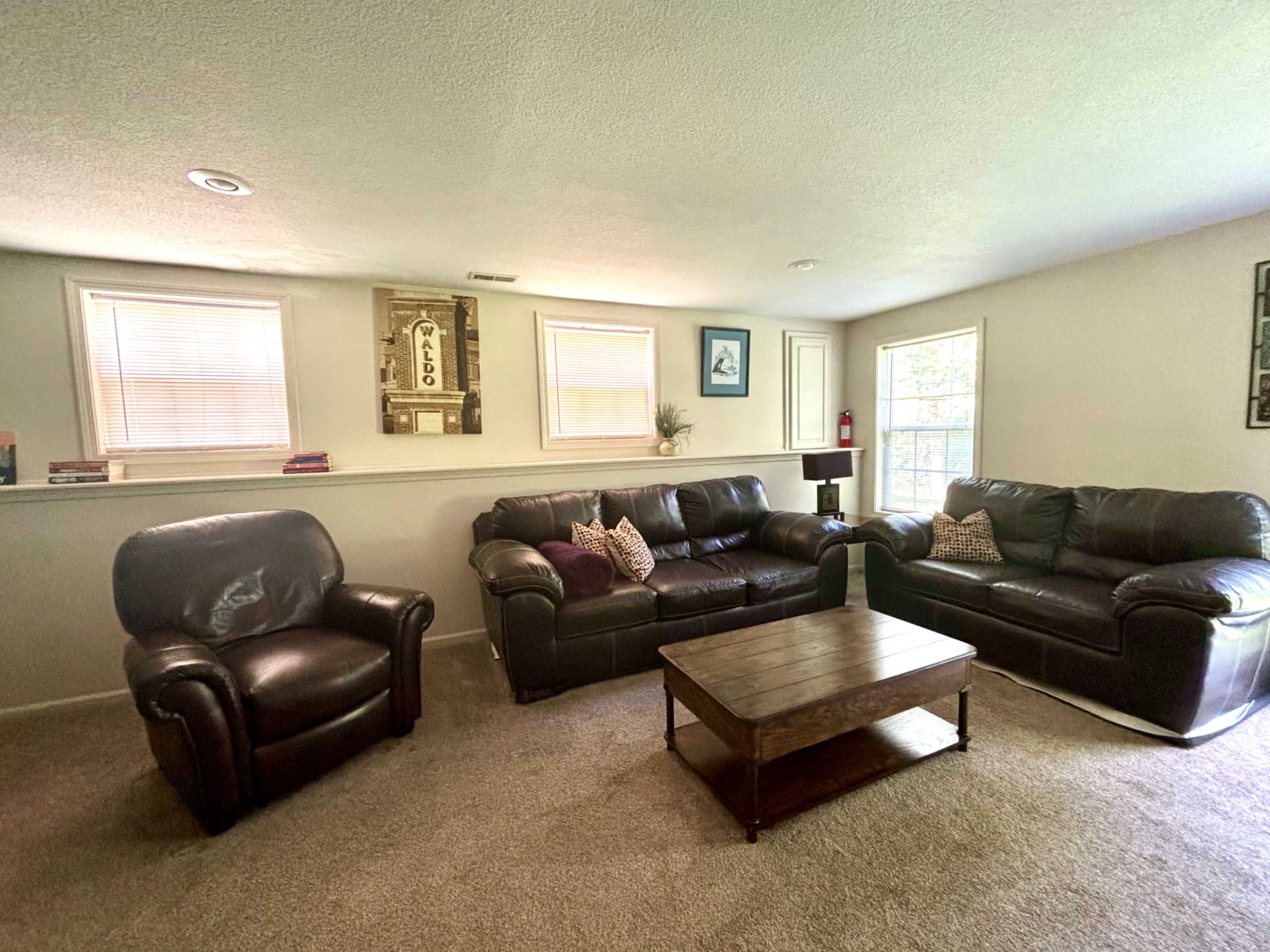 The image shows a cozy living room with two leather sofas, a reclining chair, a wooden coffee table, and large windows letting in natural light.
