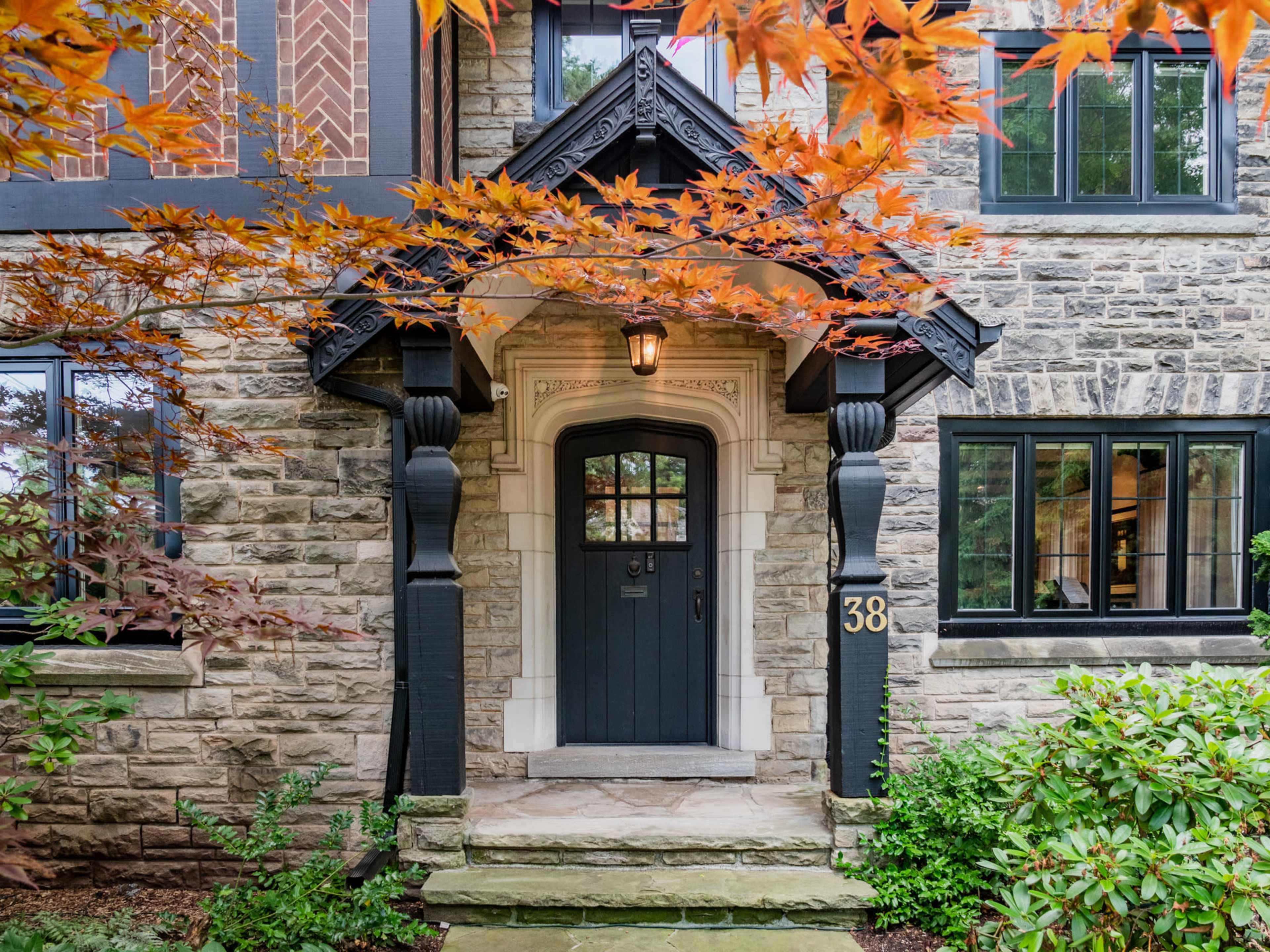 The entrance of a stone-clad house features a dark door framed by ornate woodwork and flanked by lush greenery and red maple leaves.