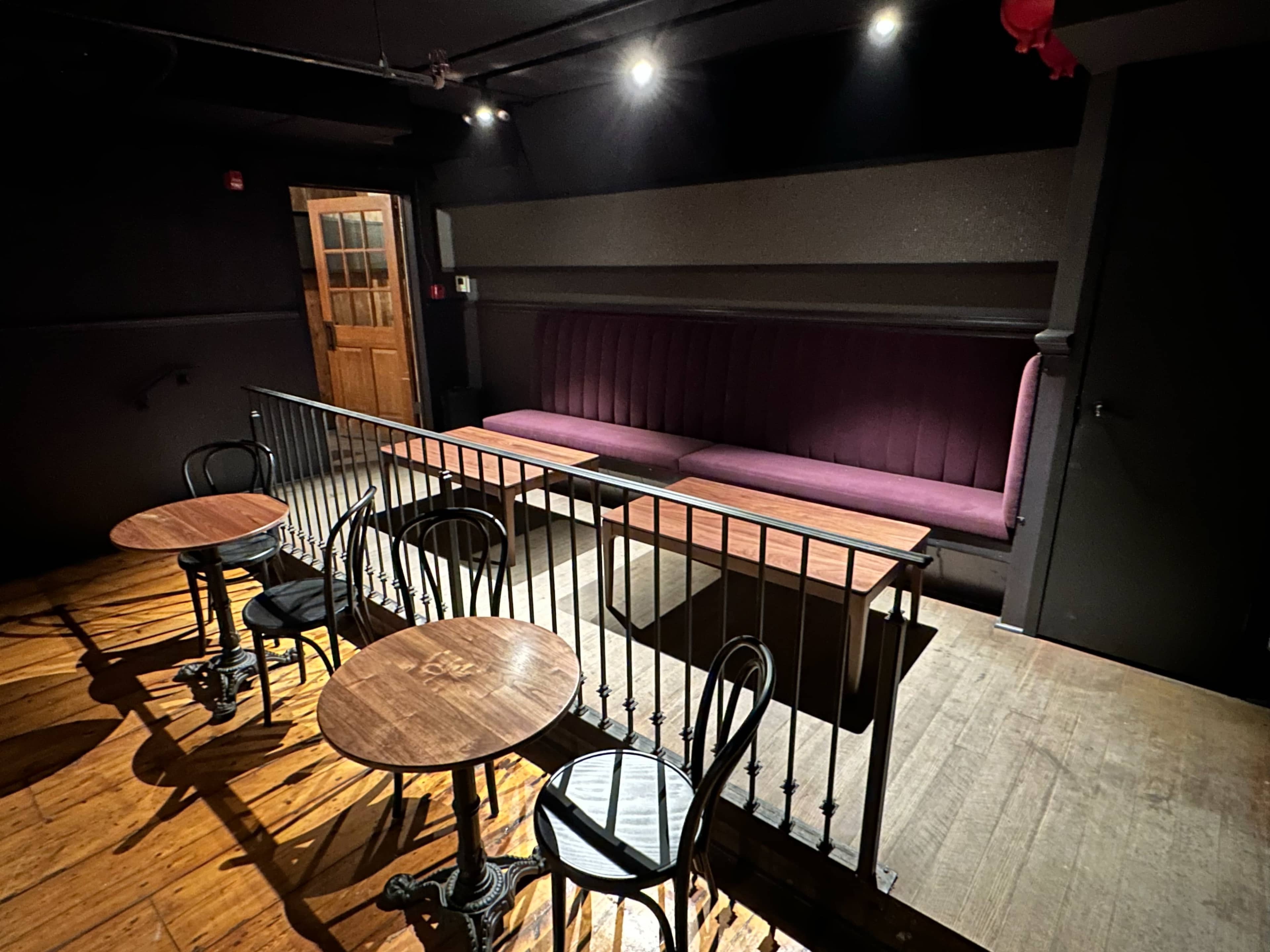 The image shows a dimly lit interior of a small venue featuring several wooden tables and chairs, along with a long purple banquette against the wall.