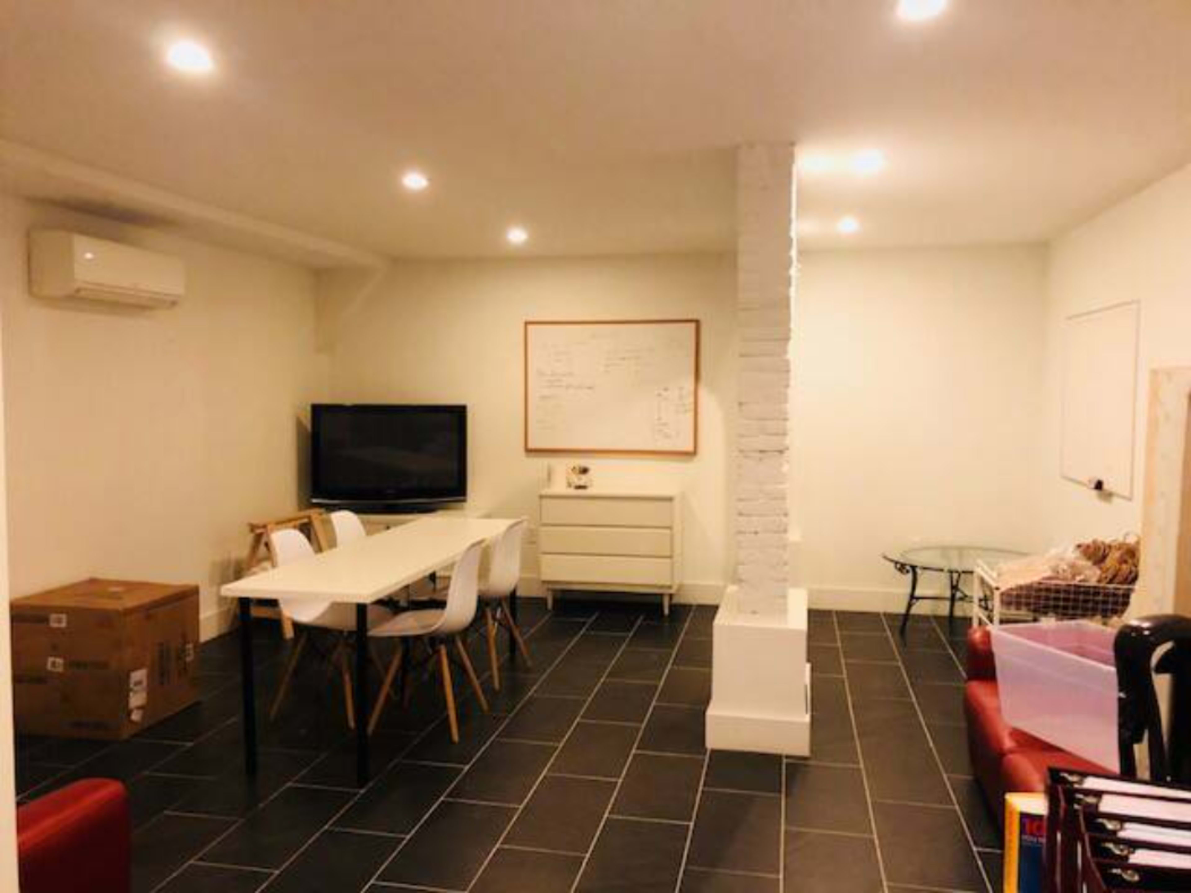 The image shows a sparsely furnished room with a whiteboard, a television, a dining table with chairs, and a glass coffee table.