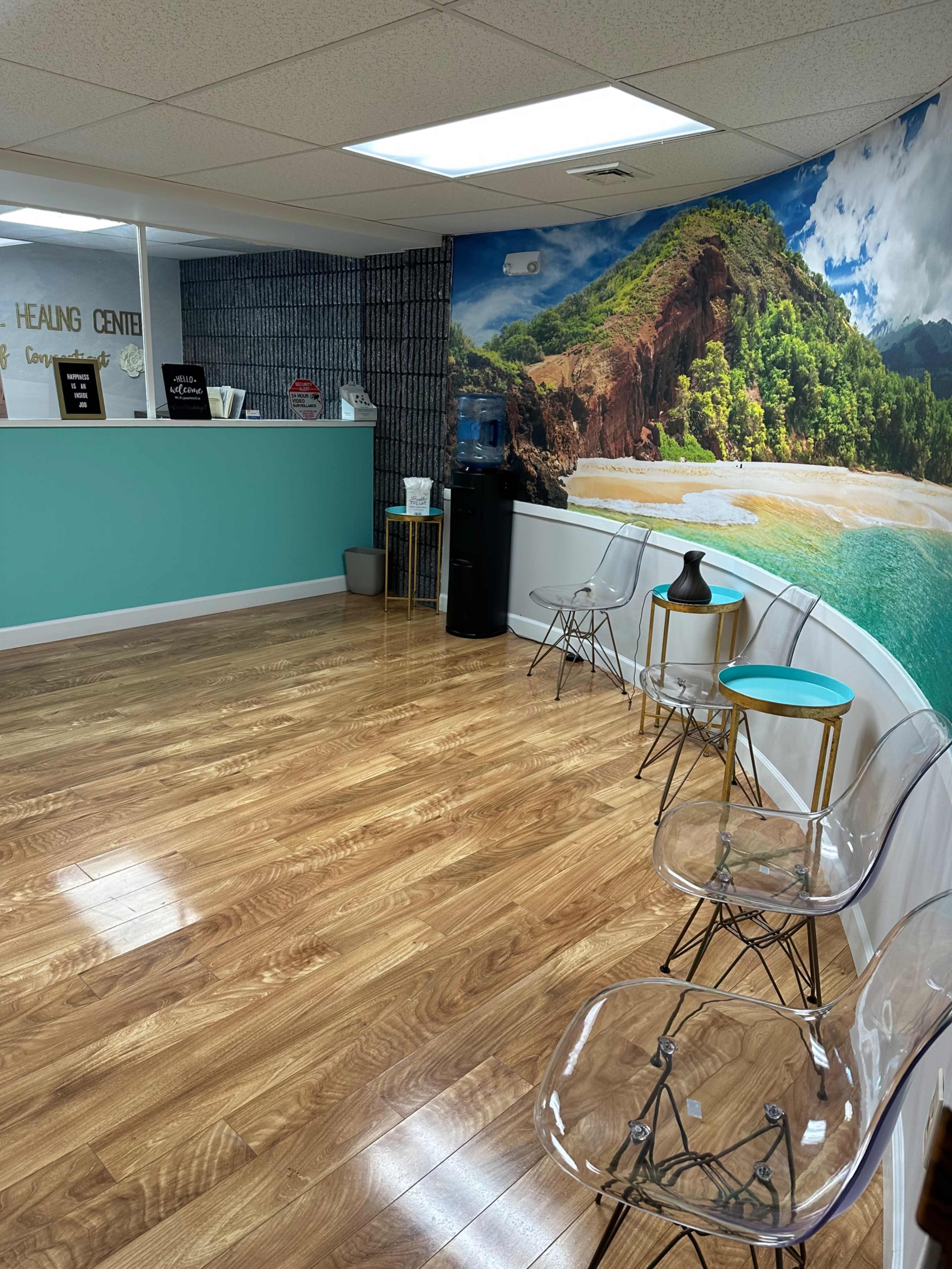 A waiting area with clear chairs, a side table, and a tropical beach mural on the wall.