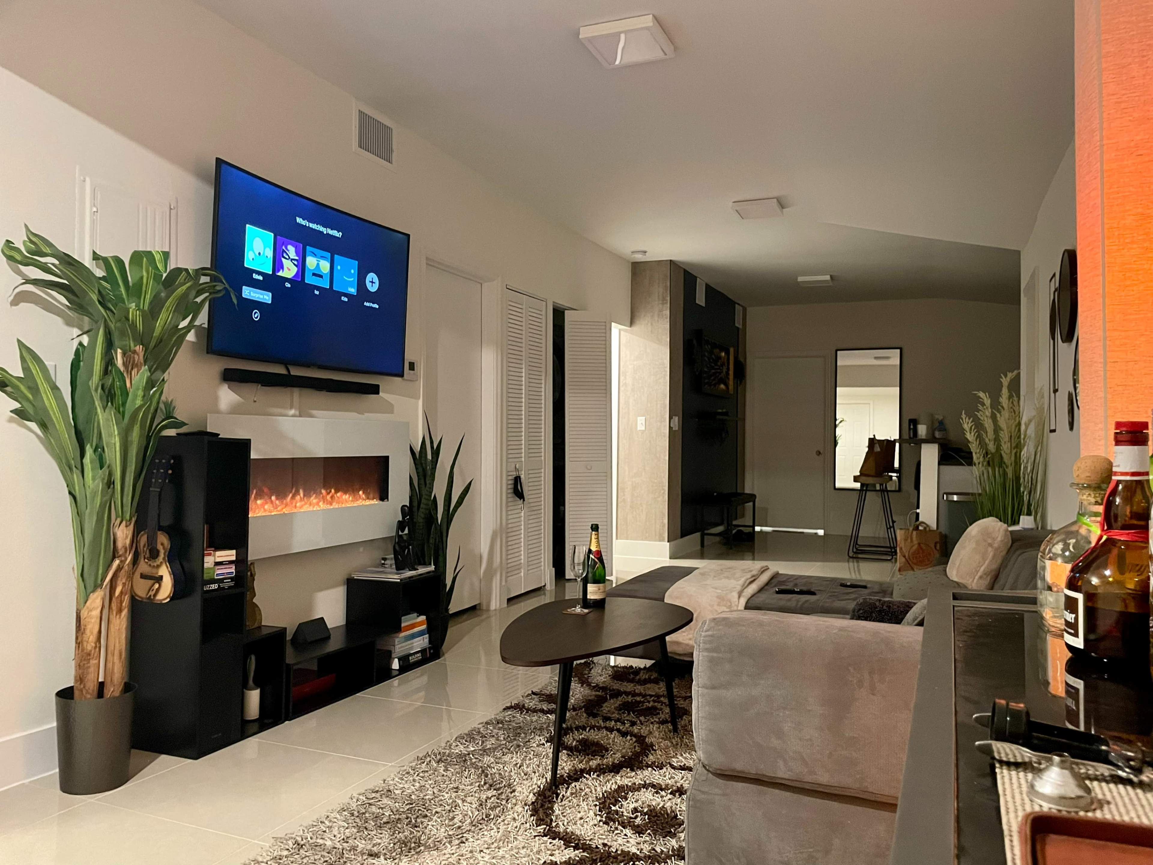 A modern living room with a wall-mounted television, a faux fireplace, a coffee table, and plush seating, featuring decorative plants and a view into an adjoining room.