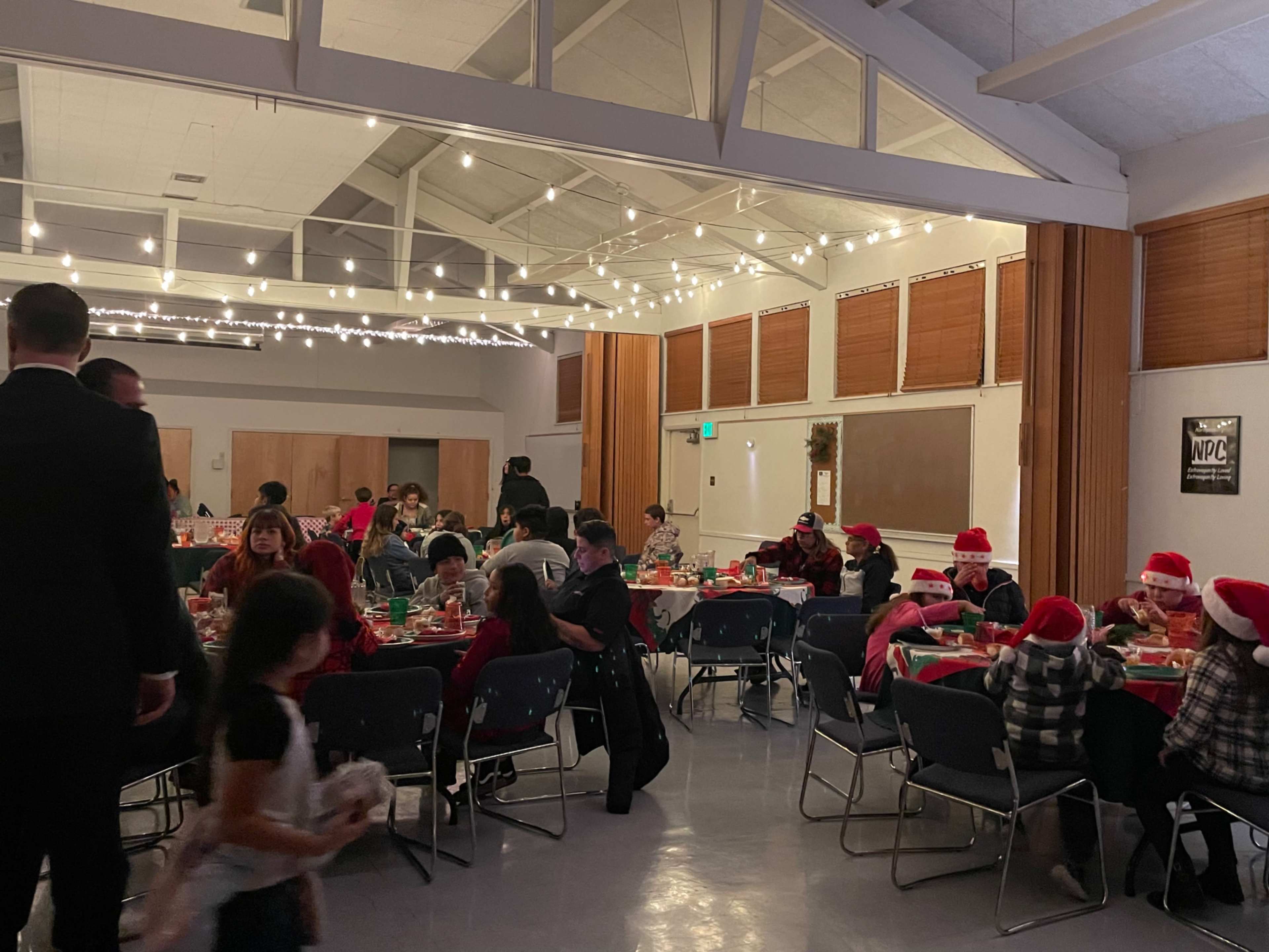 A group of people wearing Christmas hats gathers around tables decorated for a holiday party, with string lights illuminating the room.