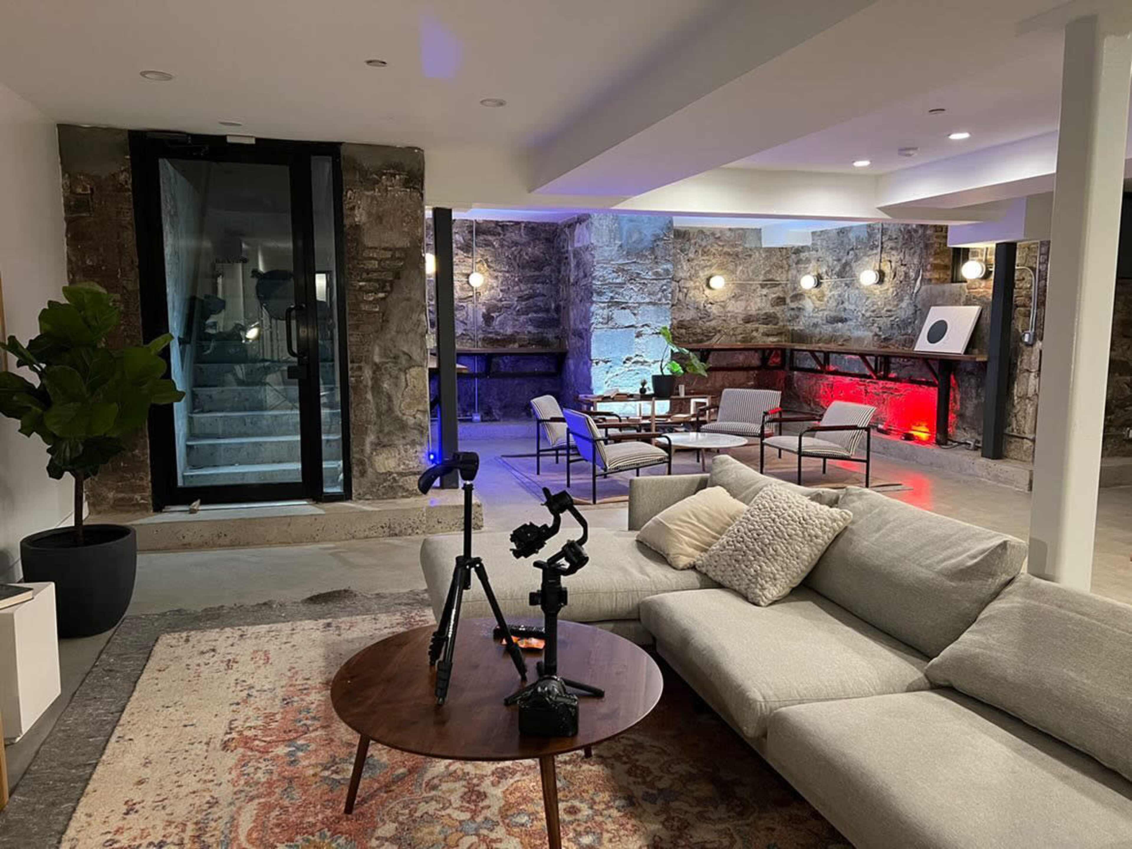 A modern basement living area features a gray sectional sofa, a round wooden coffee table, various seating arrangements, and exposed stone walls illuminated by colored lights.